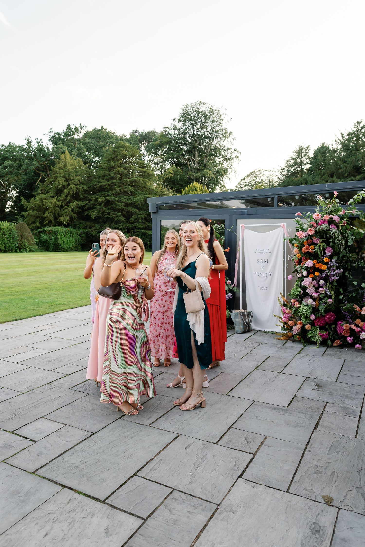 A group in colorful dresses poses together on a stone patio near a floral arch during an outdoor event.