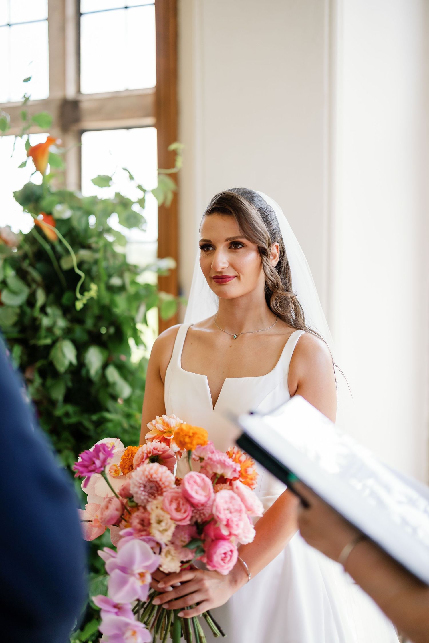 A bride in a white dress holds a vibrant pink and coral bouquet while getting ready on her wedding day.