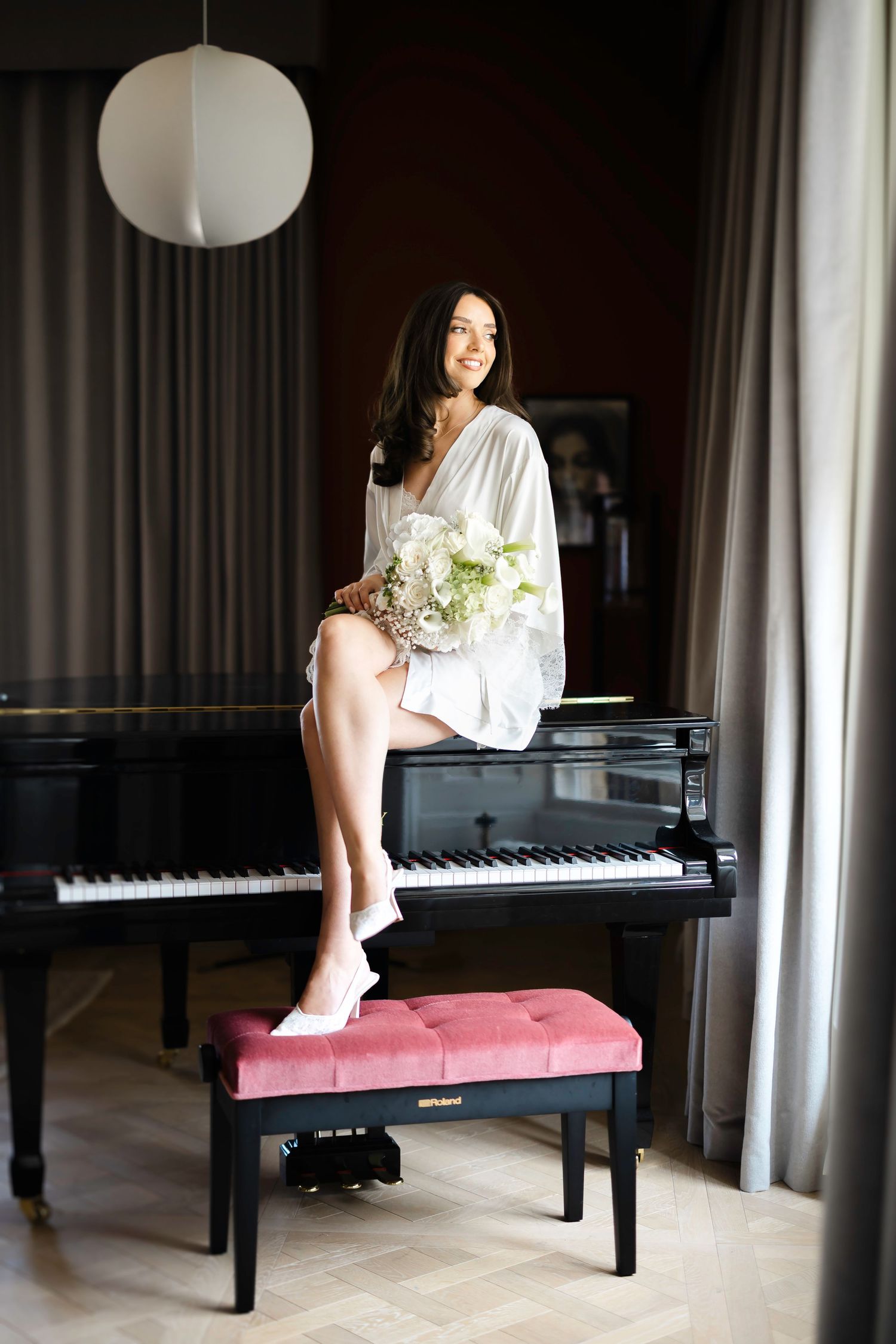 Smiling bride in white robe sits atop a black grand piano holding a bouquet of white flowers in an elegant room.