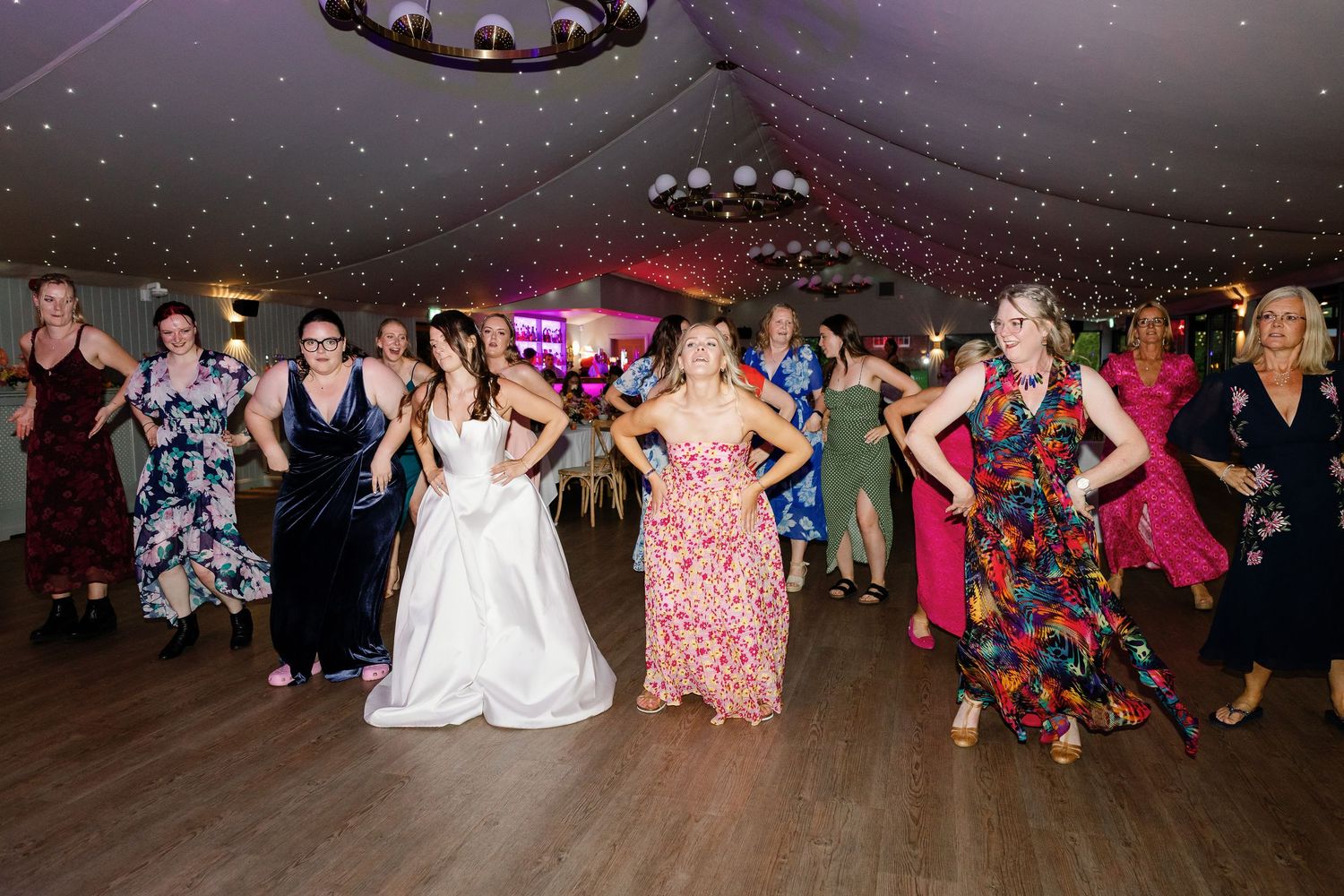 A group of people dancing together at a wedding reception under a decorated ceiling with festive lighting.