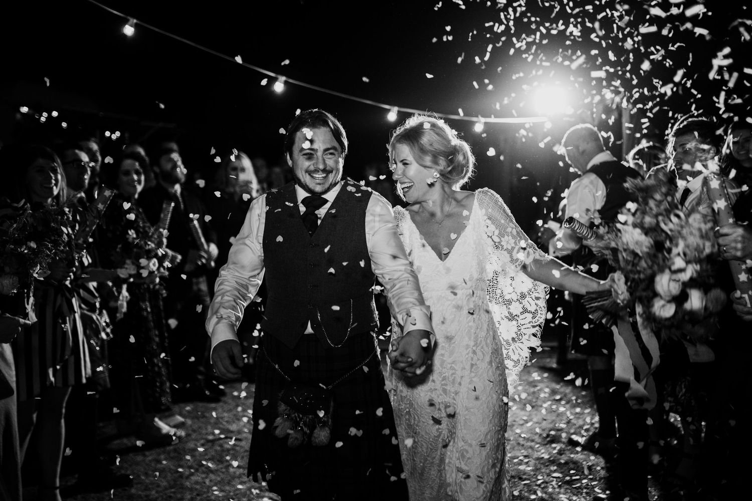 A black and white wedding photo showing a couple walking through falling confetti under string lights at night.