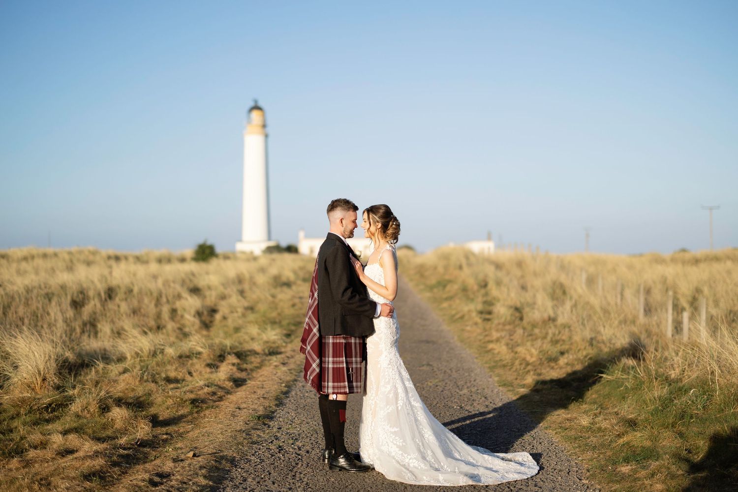 Scottish wedding couple embraces on path leading to coastal lighthouse in traditional highland attire at Broxmouth Courtyard