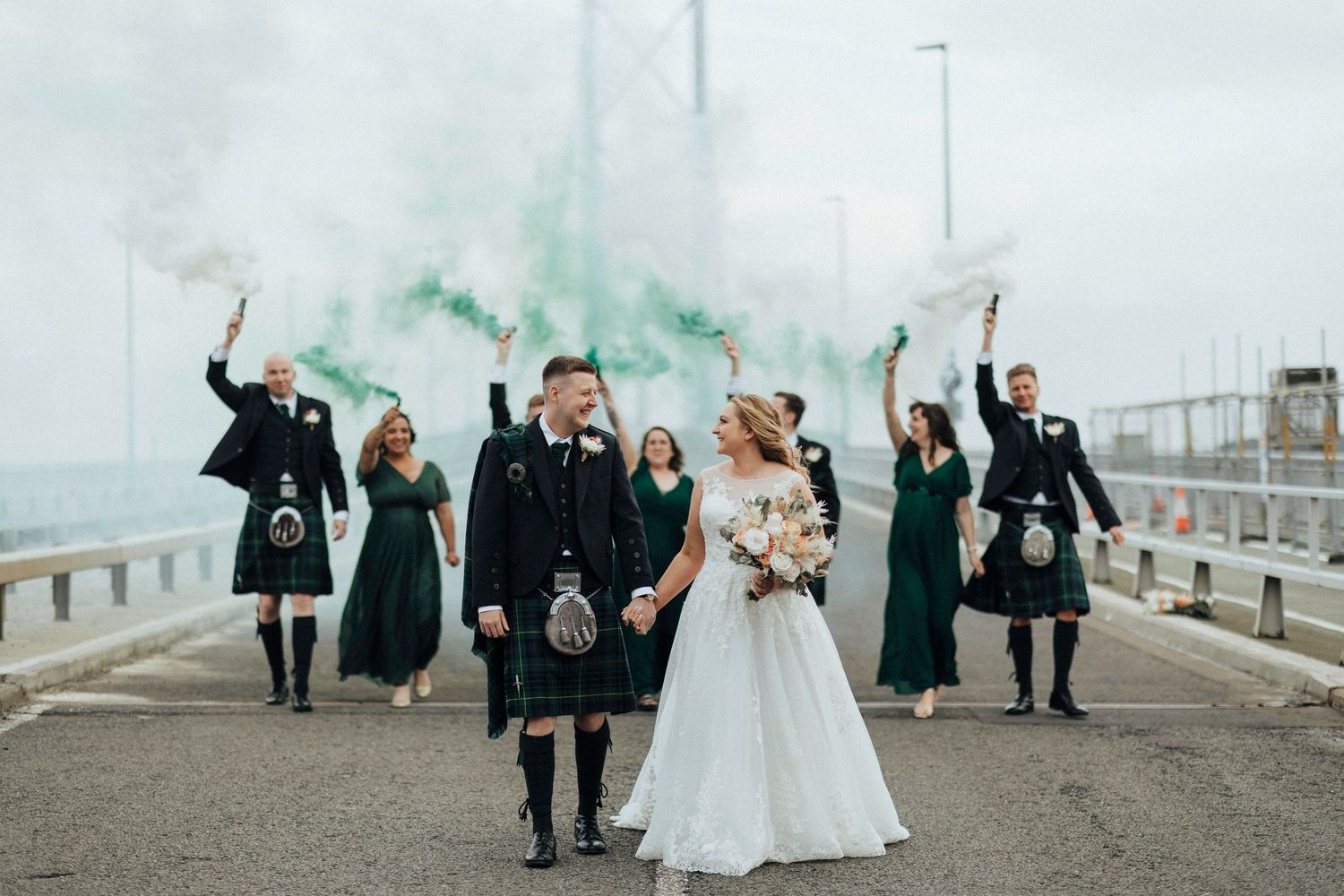 A wedding party in Scottish attire celebrating on a pier with emerald green smoke effects in the background.