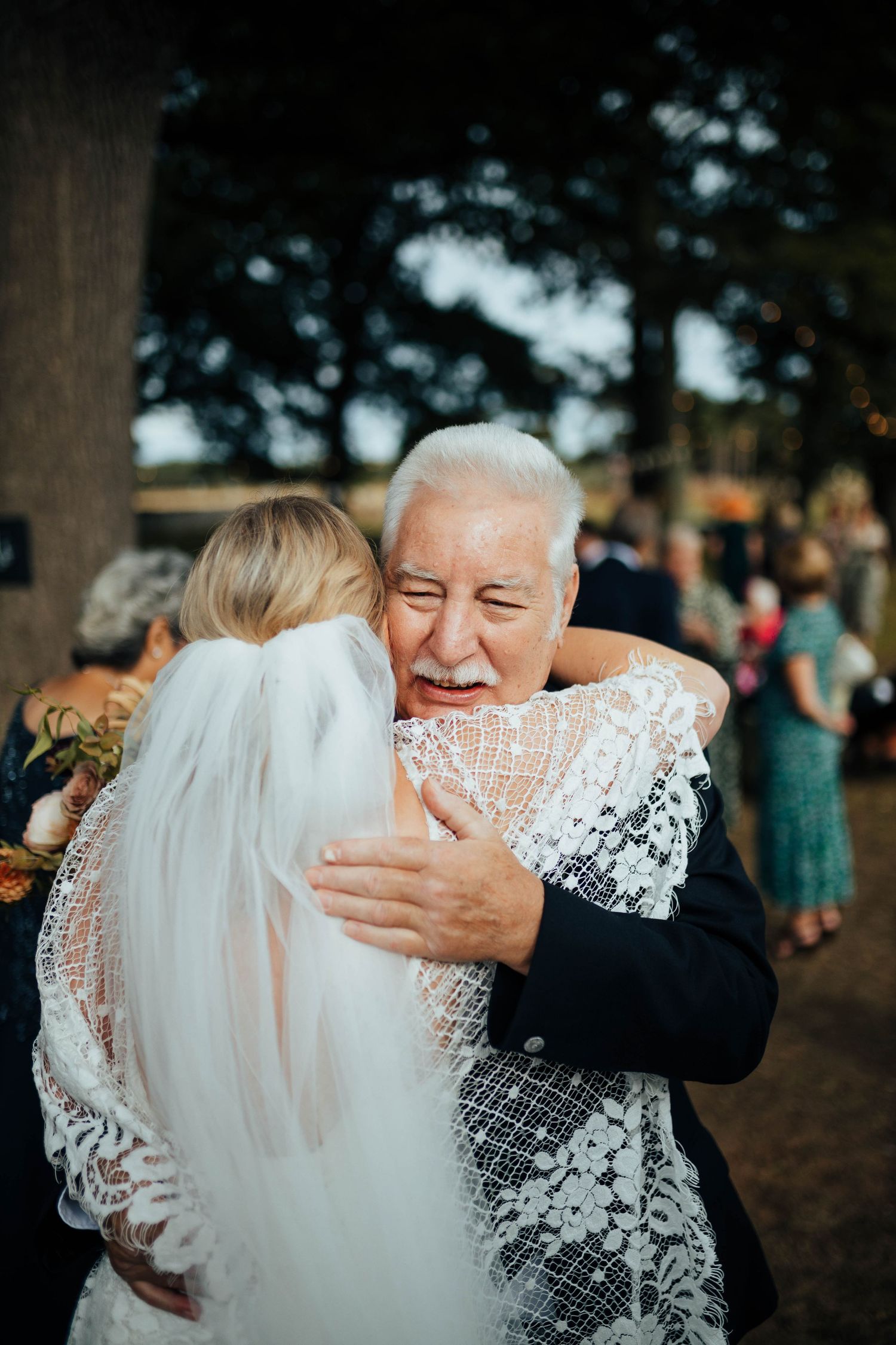 A tender embrace shared between the bride in her white lace veil and dress and an older gentleman at a wedding reception.