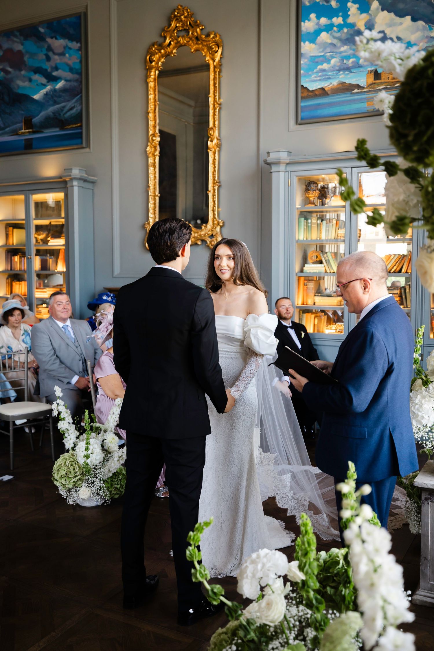 Bride and groom exchange vows indoors, surrounded by white florals, bookshelves, and a gold mirror during elegant wedding ceremony.