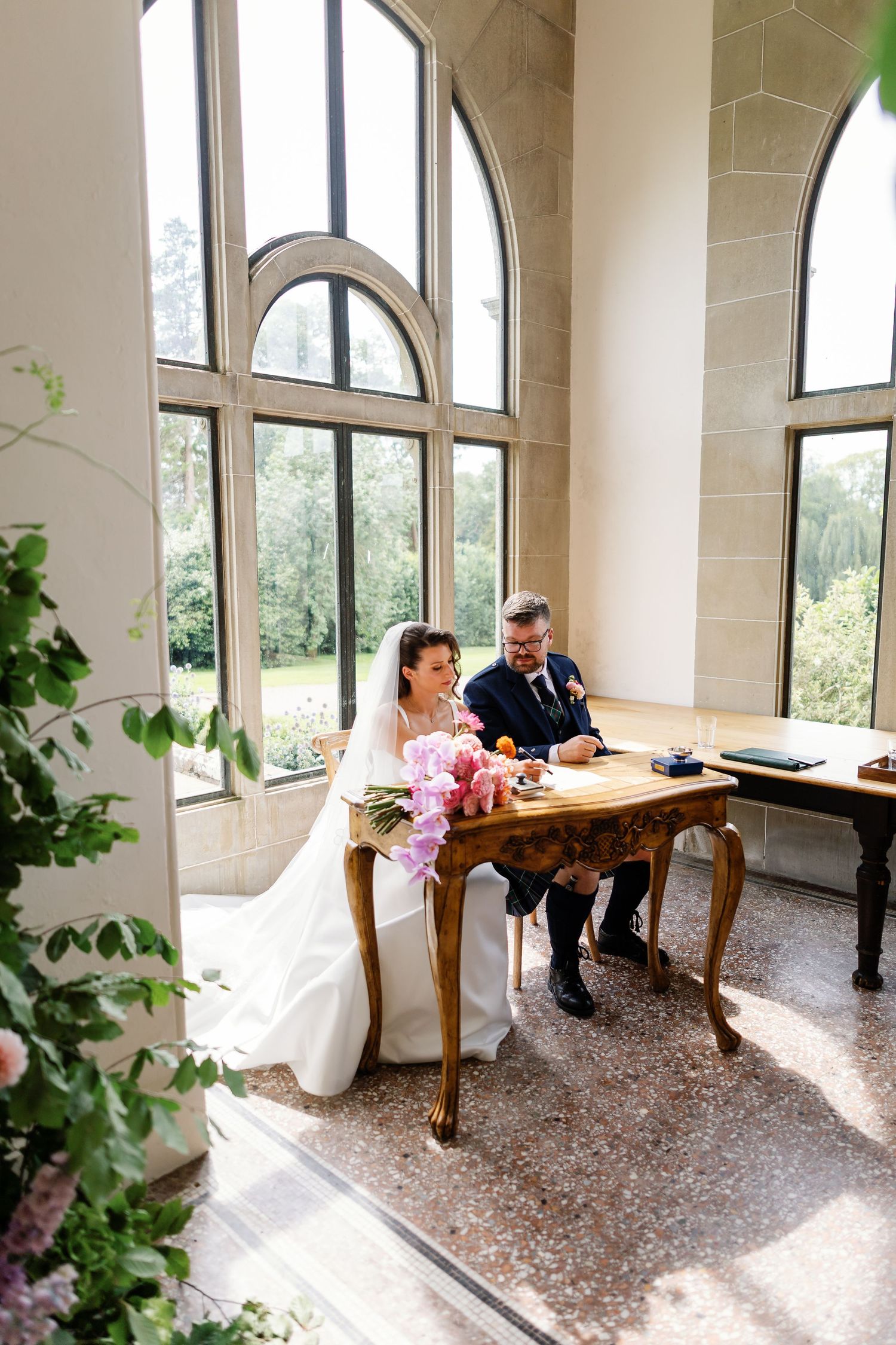 Elegant sunlit room with arched windows and ornate gold table where a couple sits together in formal wedding attire.