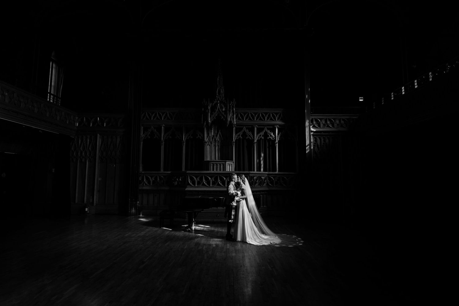 Dramatic black and white church photo shows a couple in wedding attire standing in a shaft of light amid Gothic architecture.