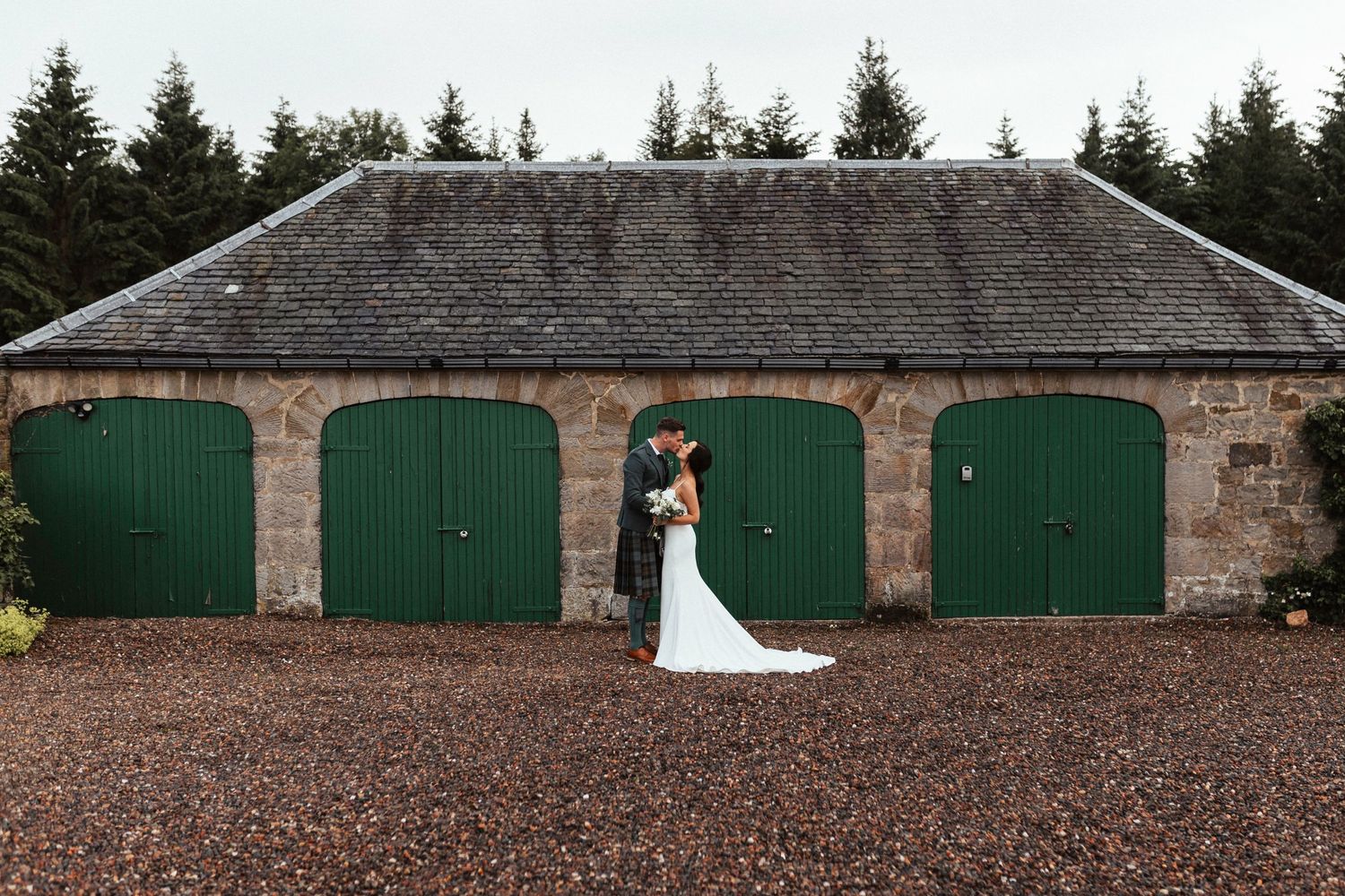 A couple embraces outside historic stone garage building with green doors and gravel courtyard in Scotland.