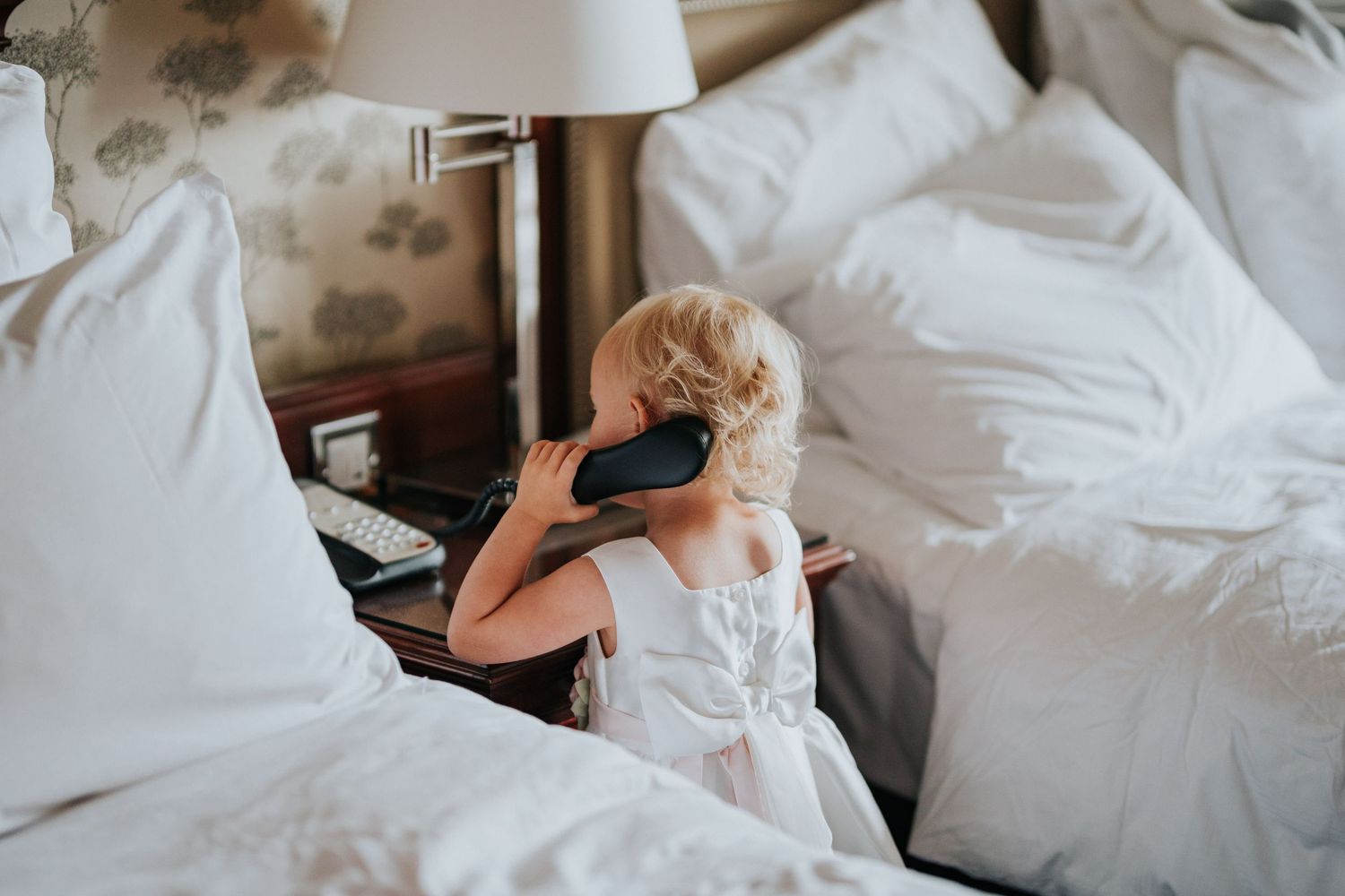 Someone in white sitting on unmade bed while talking on a phone with white bedding and table lamp visible.