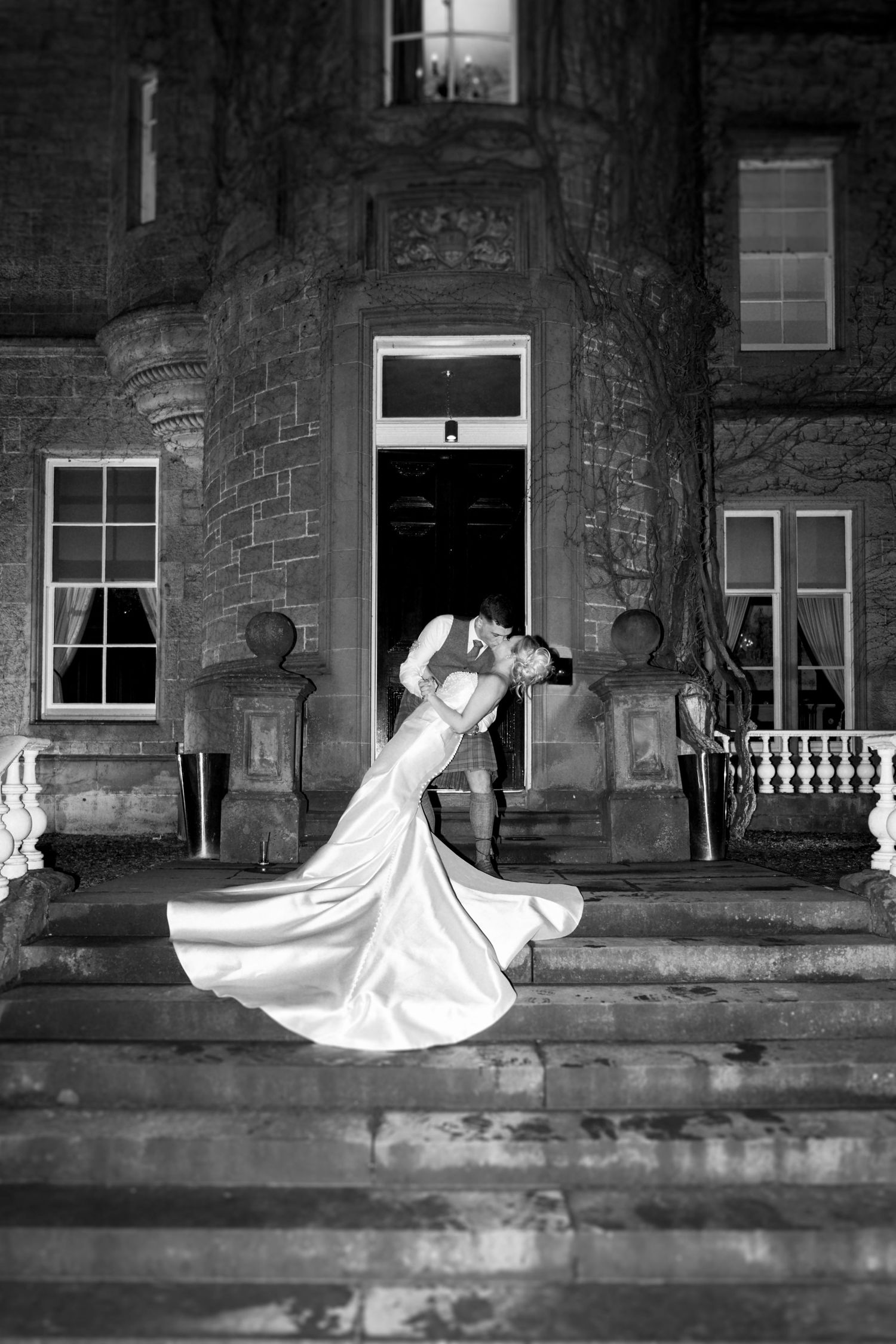 Black and white photo of bride and groom sharing a romantic dip kiss on steps of grand stone manor at night.