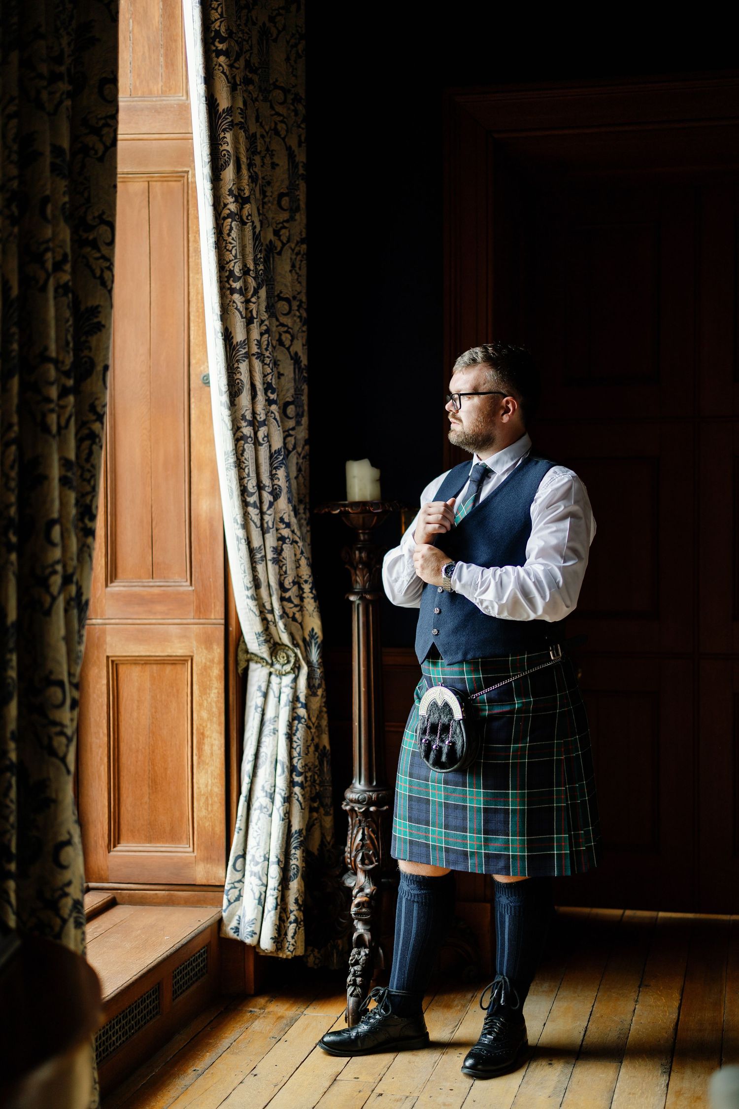 A person wearing a traditional Scottish kilt and waistcoat stands by an elegant window with ornate curtains.
