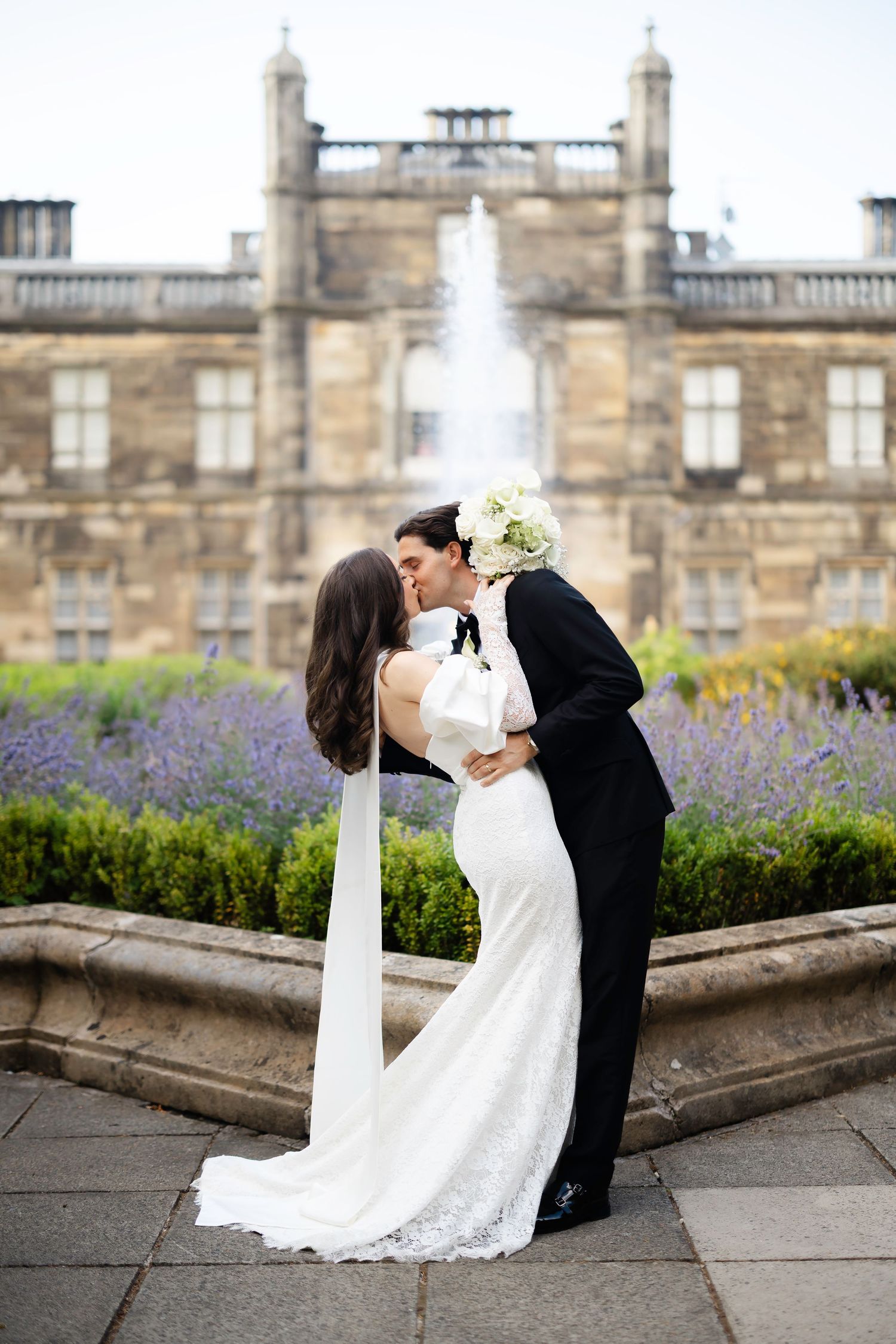 A bride and groom share a romantic kiss in a lace wedding gown and black tuxedo outside an elegant stone mansion.