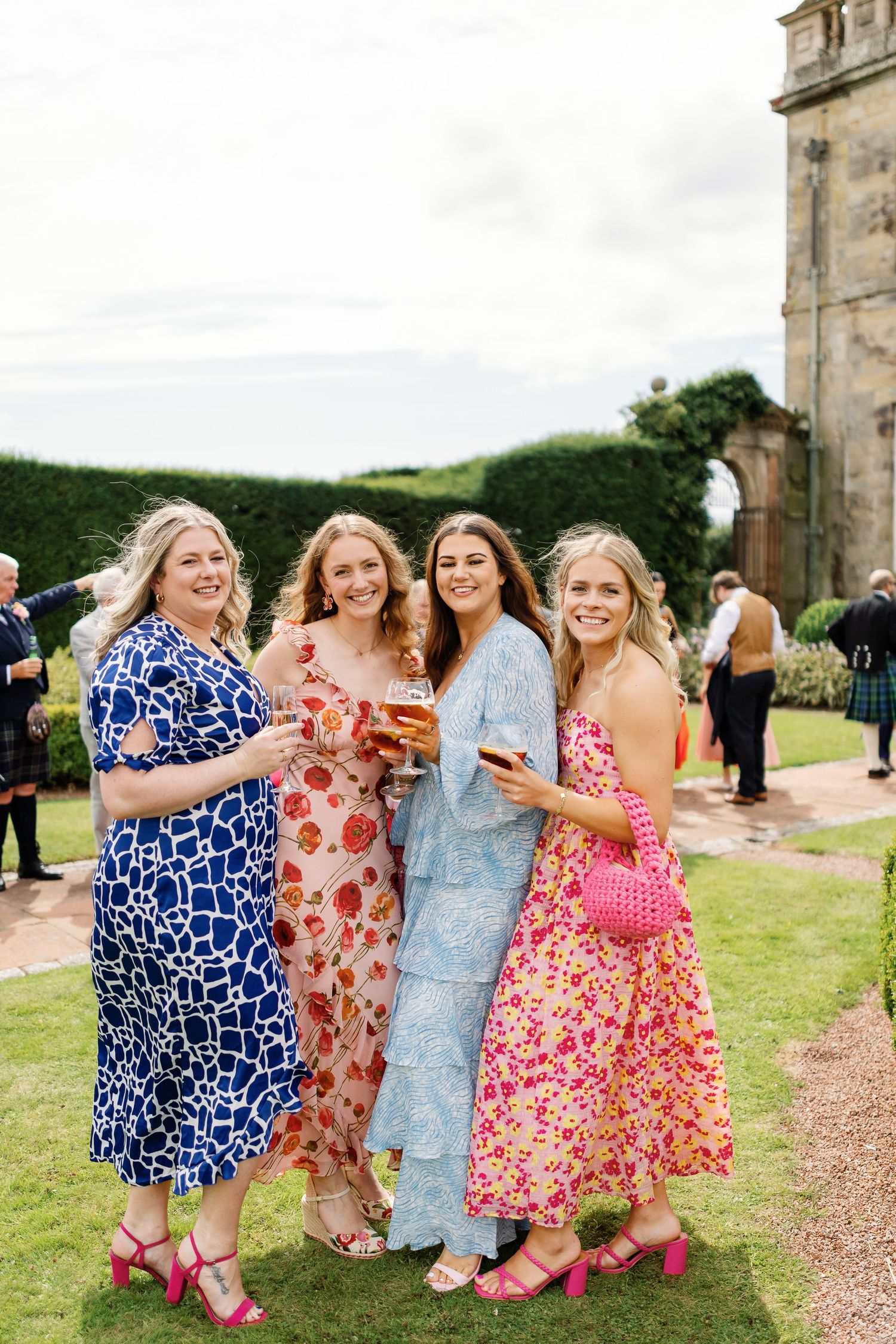 A group of wedding guests in colorful floral dresses and pink shoes laughing together on a grassy path.