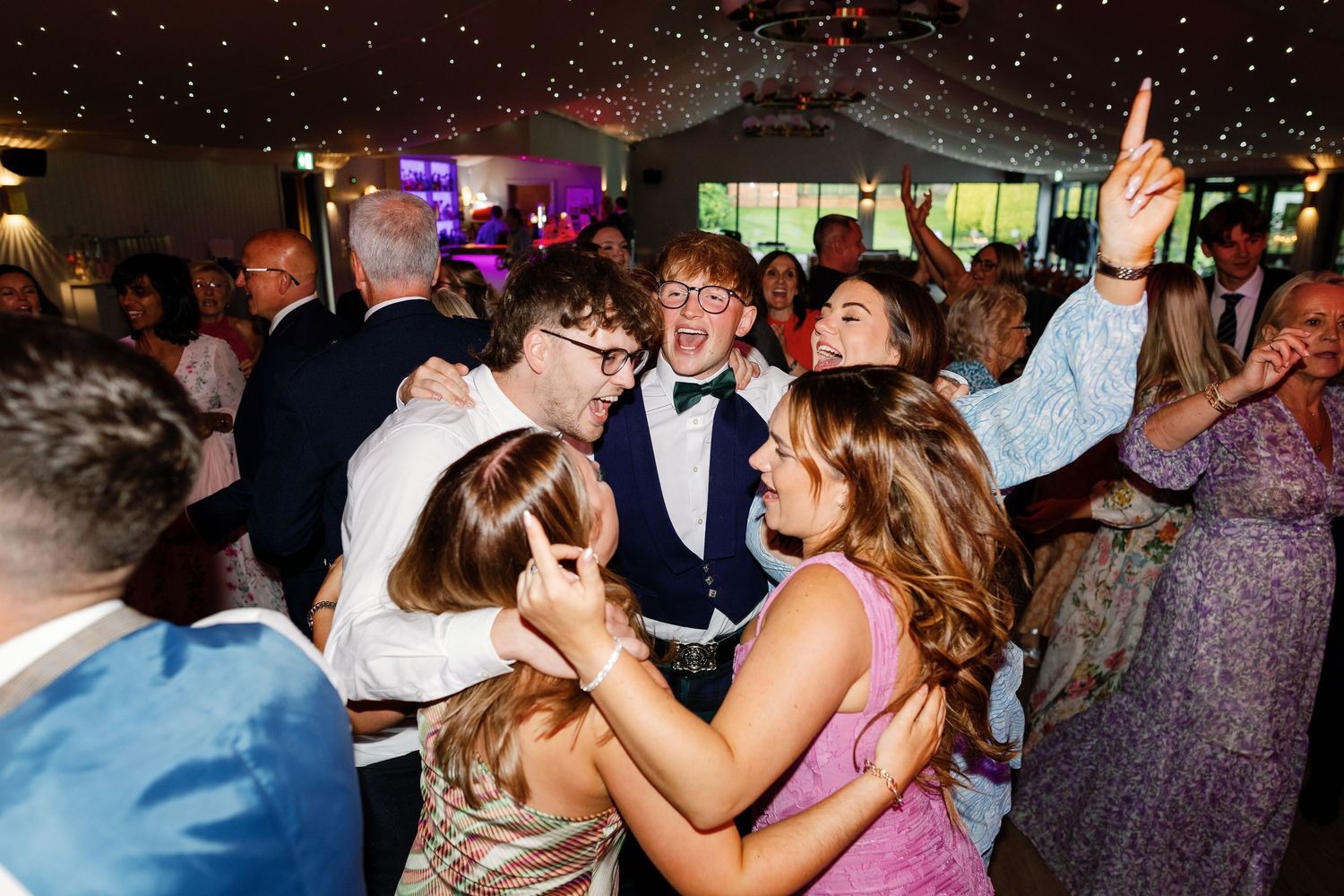 Wedding guests dance and celebrate on a crowded dance floor under colorful party lights.