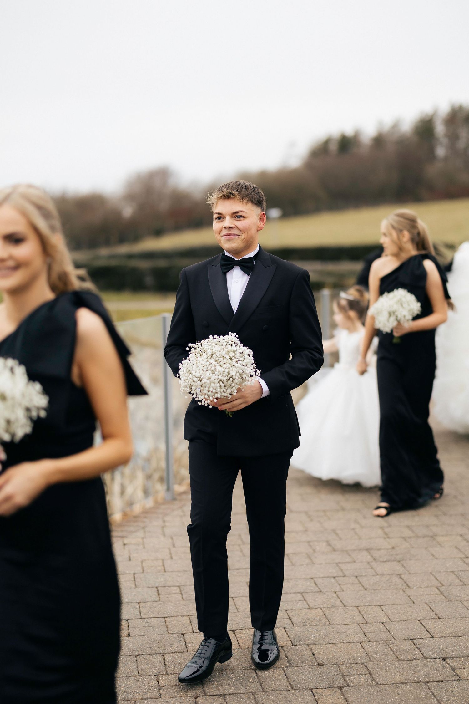 Wedding party member in black suit and bow tie holding baby's breath bouquet stands on brick patio at outdoor ceremony.