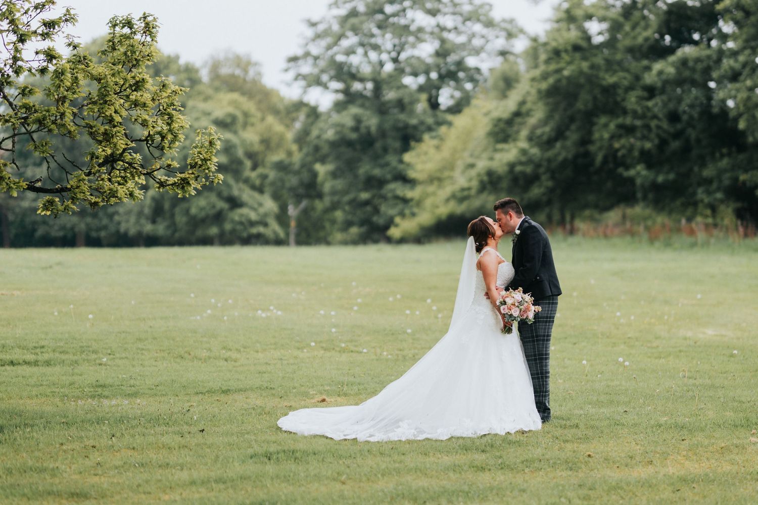A couple in wedding attire stands together in a serene green field surrounded by trees.