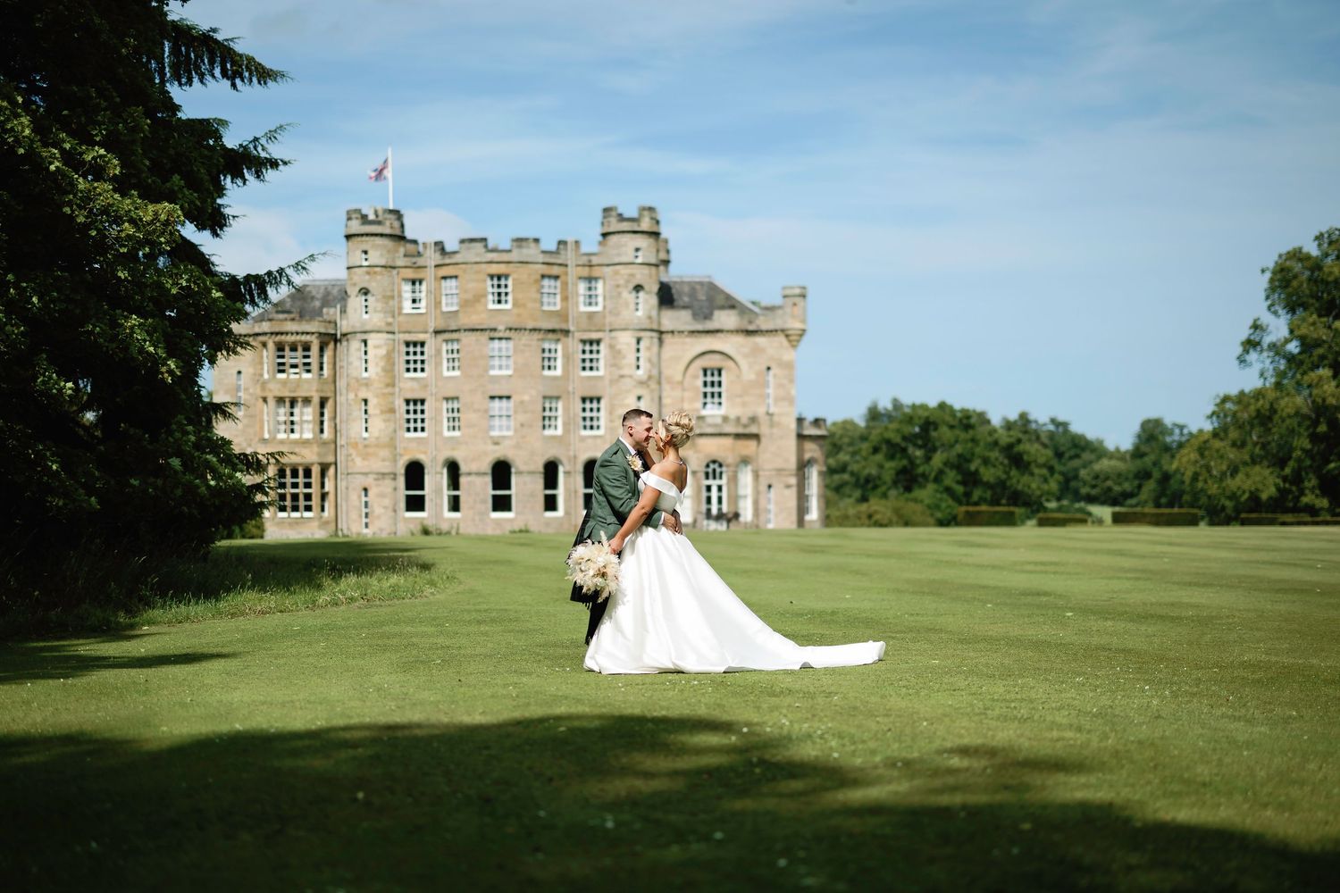 A stunning wedding couple poses on a vast green lawn in front of an elegant historic stone mansion on a sunny day.