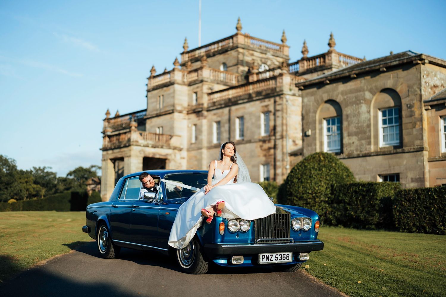 A classic blue Rolls Royce parked on the lawn of an elegant historic mansion under bright blue skies.
