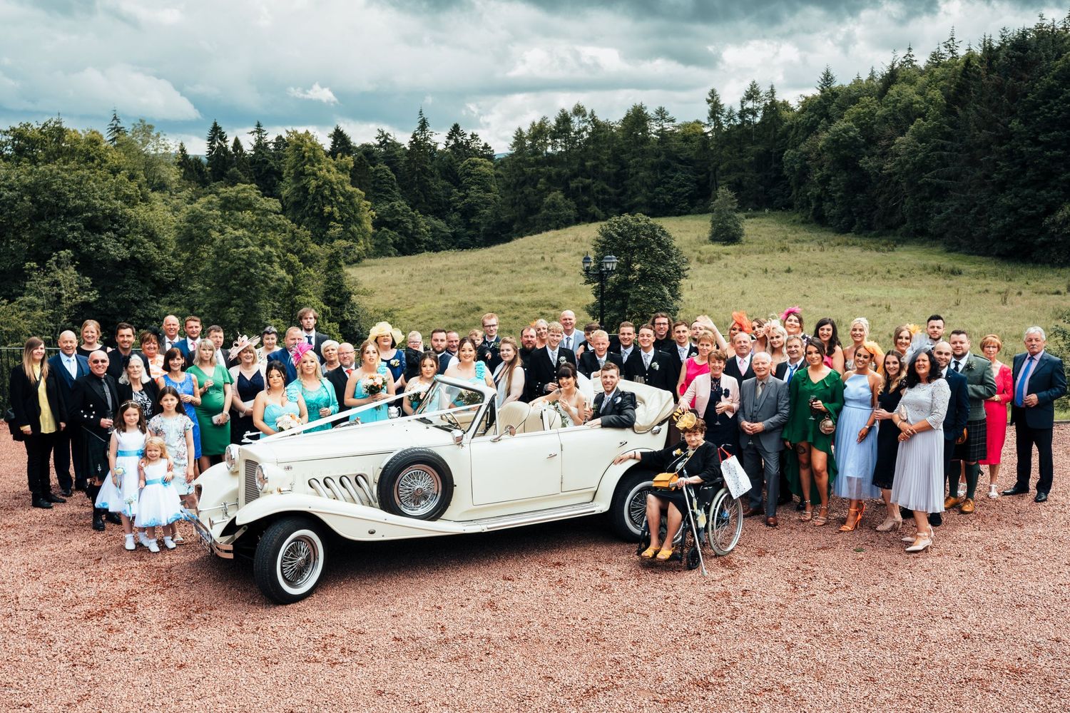 A large wedding party poses outdoors on a hillside with a vintage white luxury car.