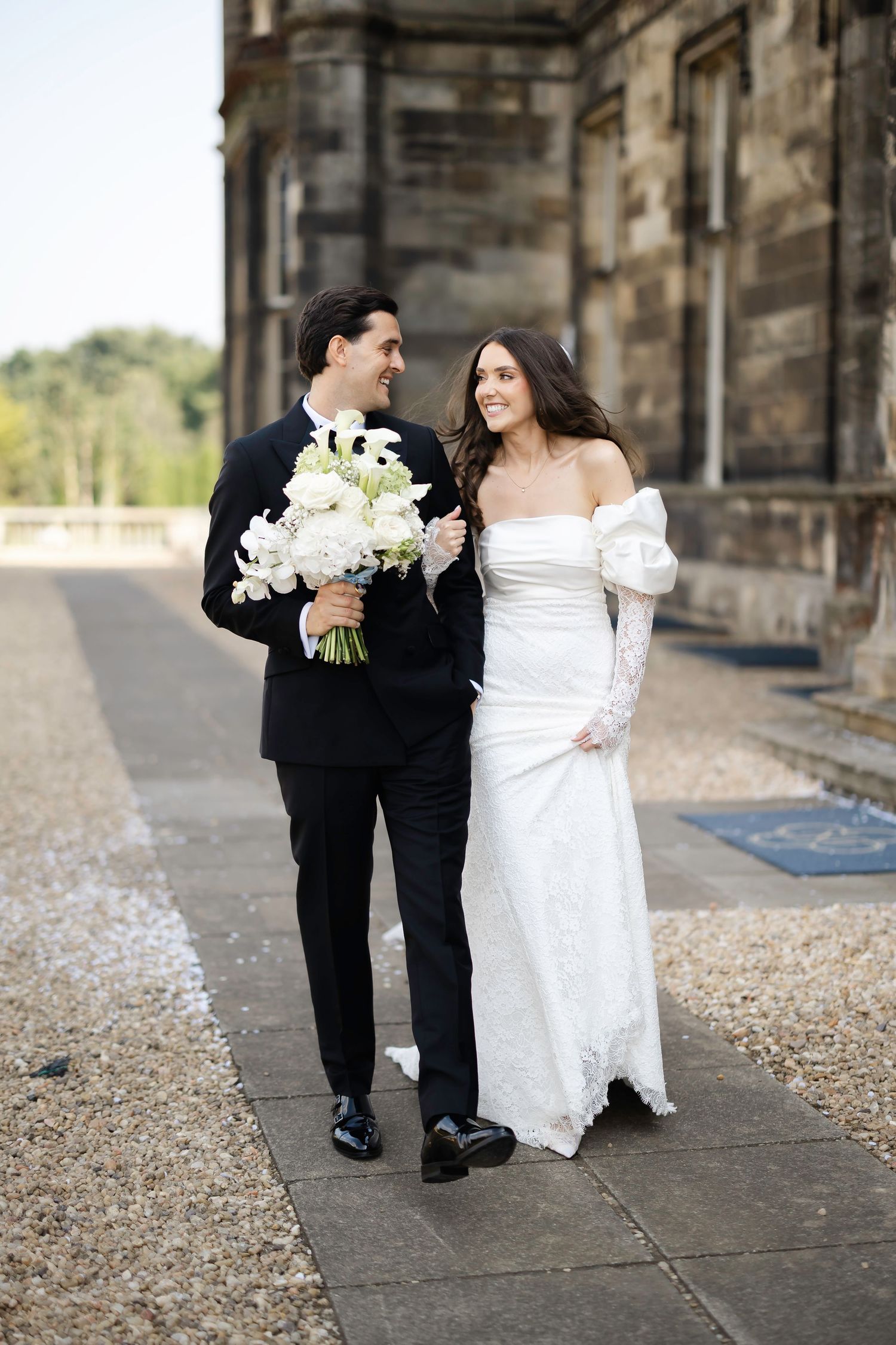 A bride and groom walk together outside a stone mansion, she in an off-shoulder white gown holding white florals.