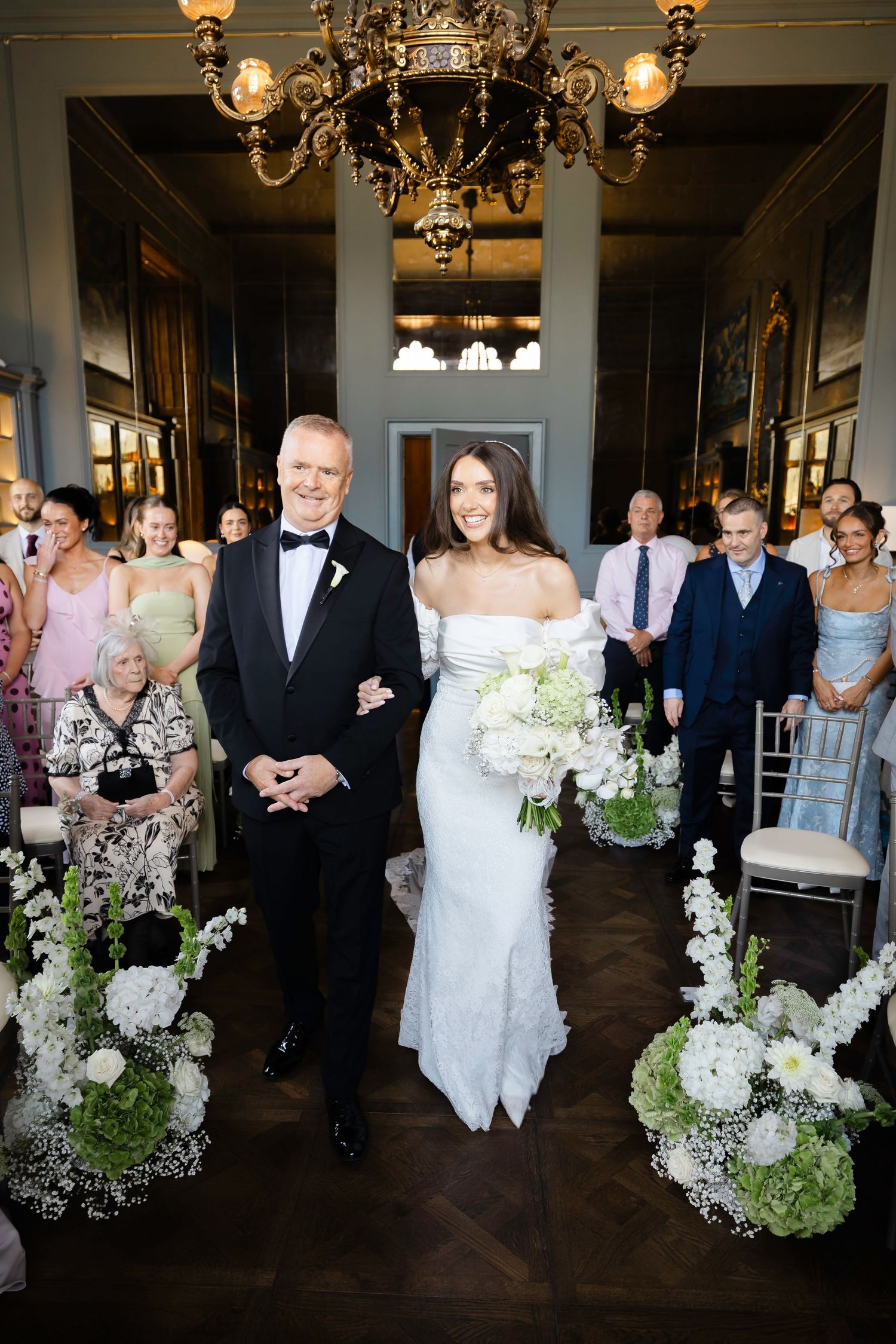 Bride in white off-shoulder gown walks the aisle with escort amid white floral arrangements in elegant chandelier-lit venue.