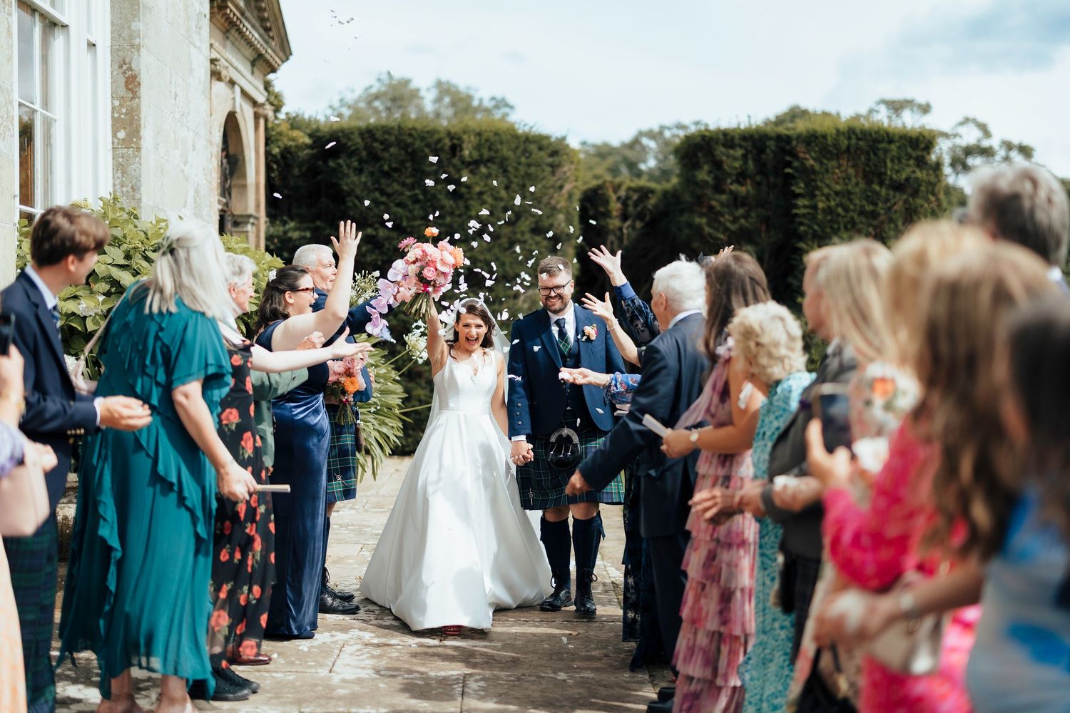 Wedding guests toss confetti over the newlyweds in celebration outside a stone venue as they exit as a married couple.