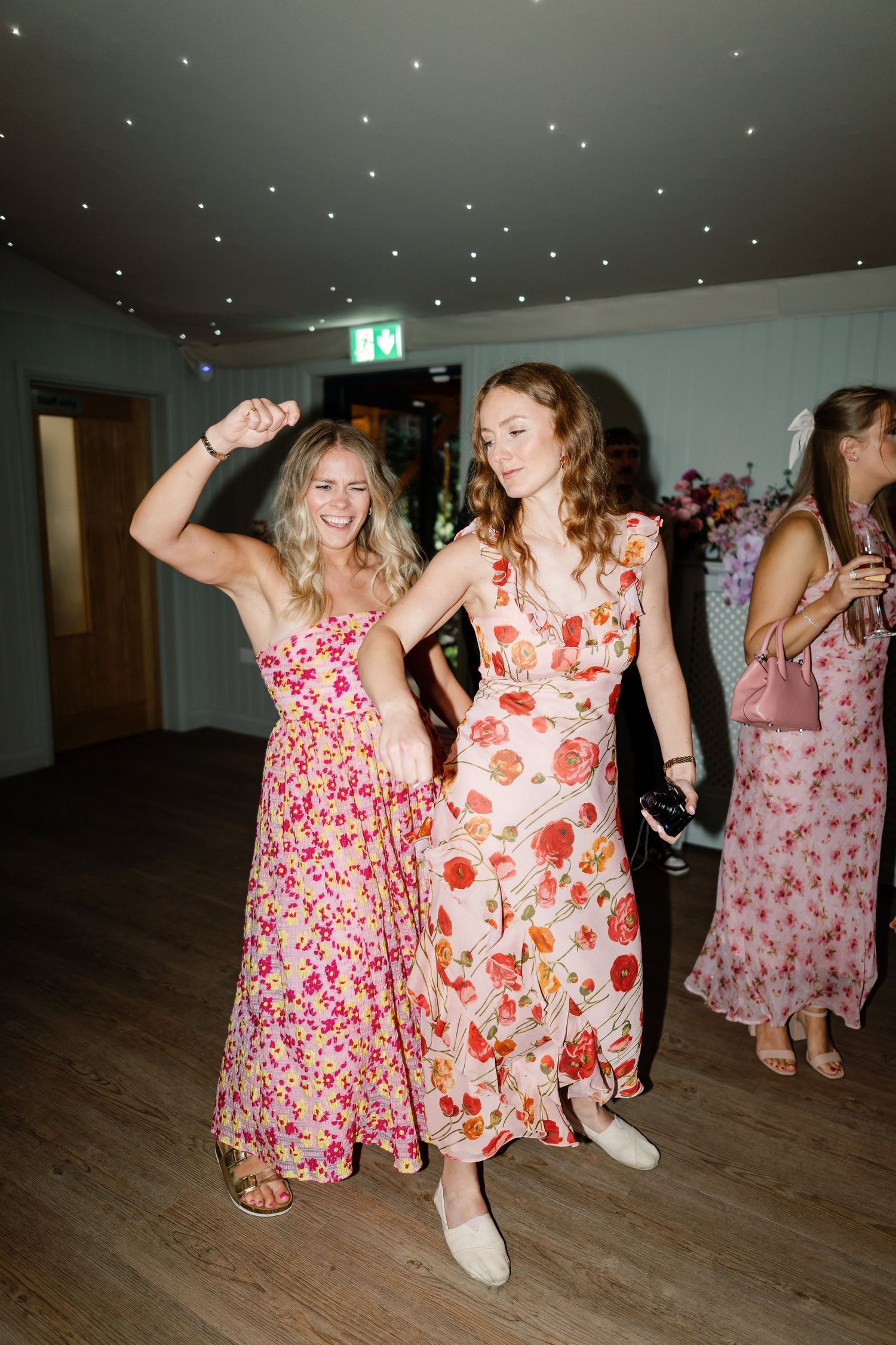 People in floral dresses dance together on a wooden floor during what appears to be a celebration or party.