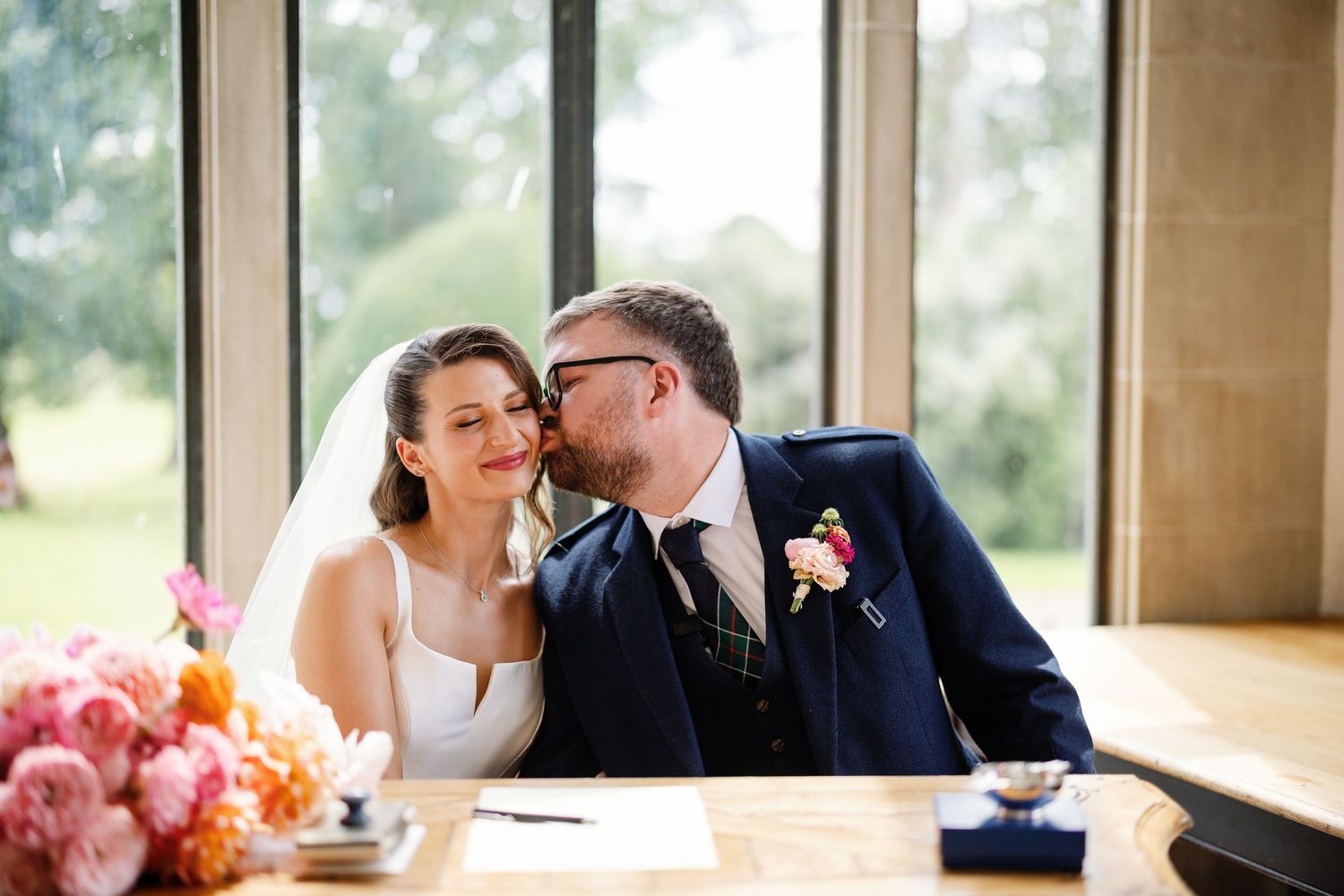 A couple shares an intimate moment near large windows in a bright room with pink flowers in the background.