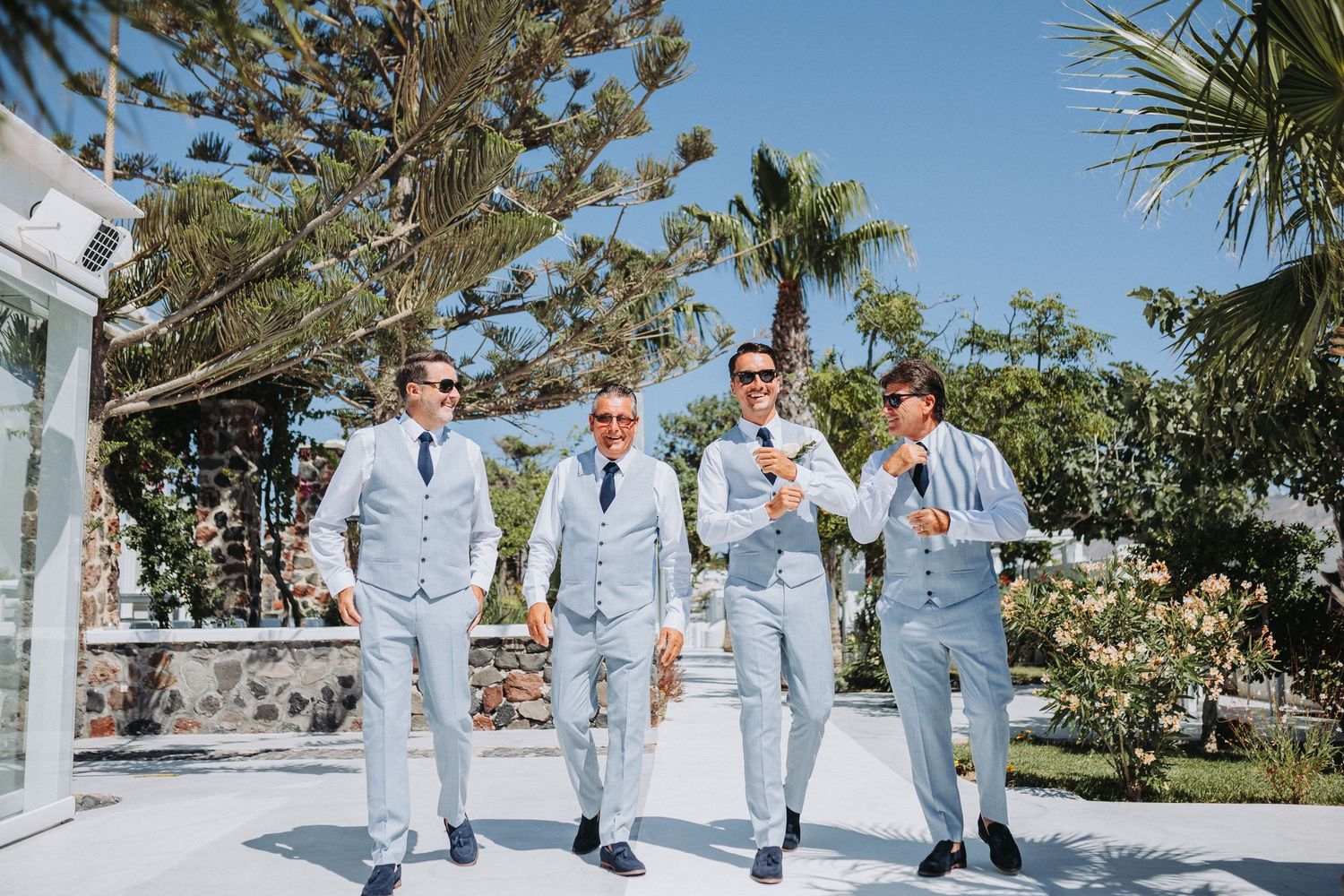 Four people wearing matching light gray suits walk together on a sunny path lined with tropical palm trees and greenery.