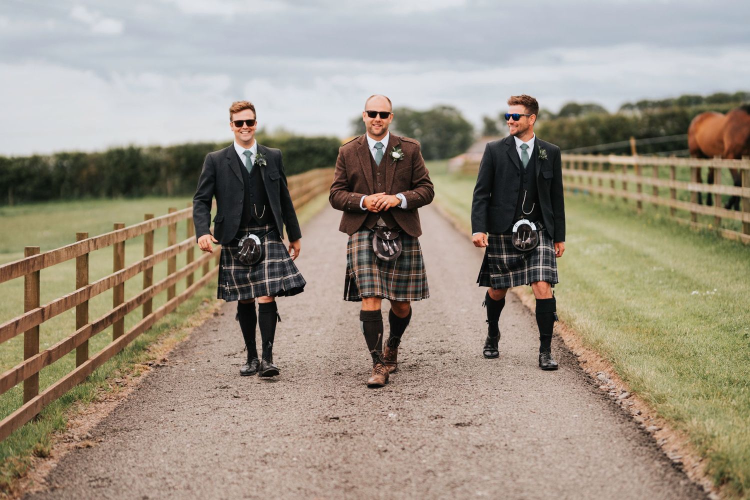 Three people in traditional Scottish kilts and formal attire walk together on a path lined with wooden fencing.