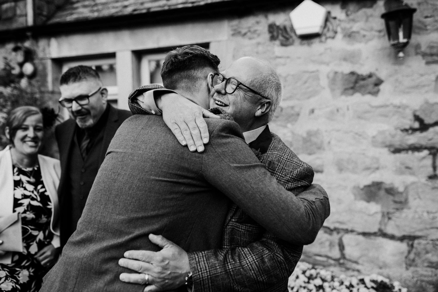 Two men in vintage clothing share a warm embrace against a stone wall while others look on in black and white.