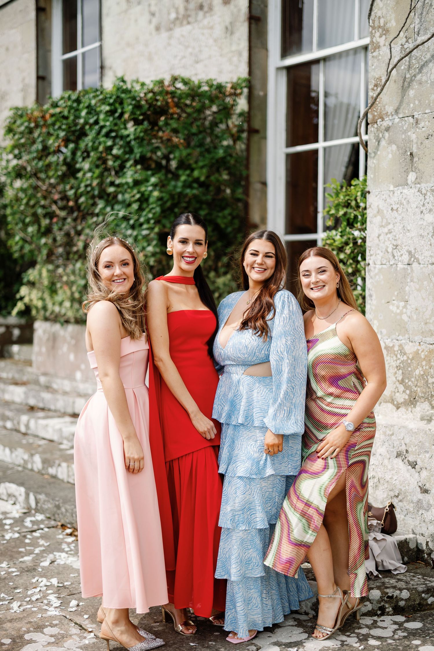 A group of friends in colorful dresses laugh and pose together against a stone wall with climbing vines.