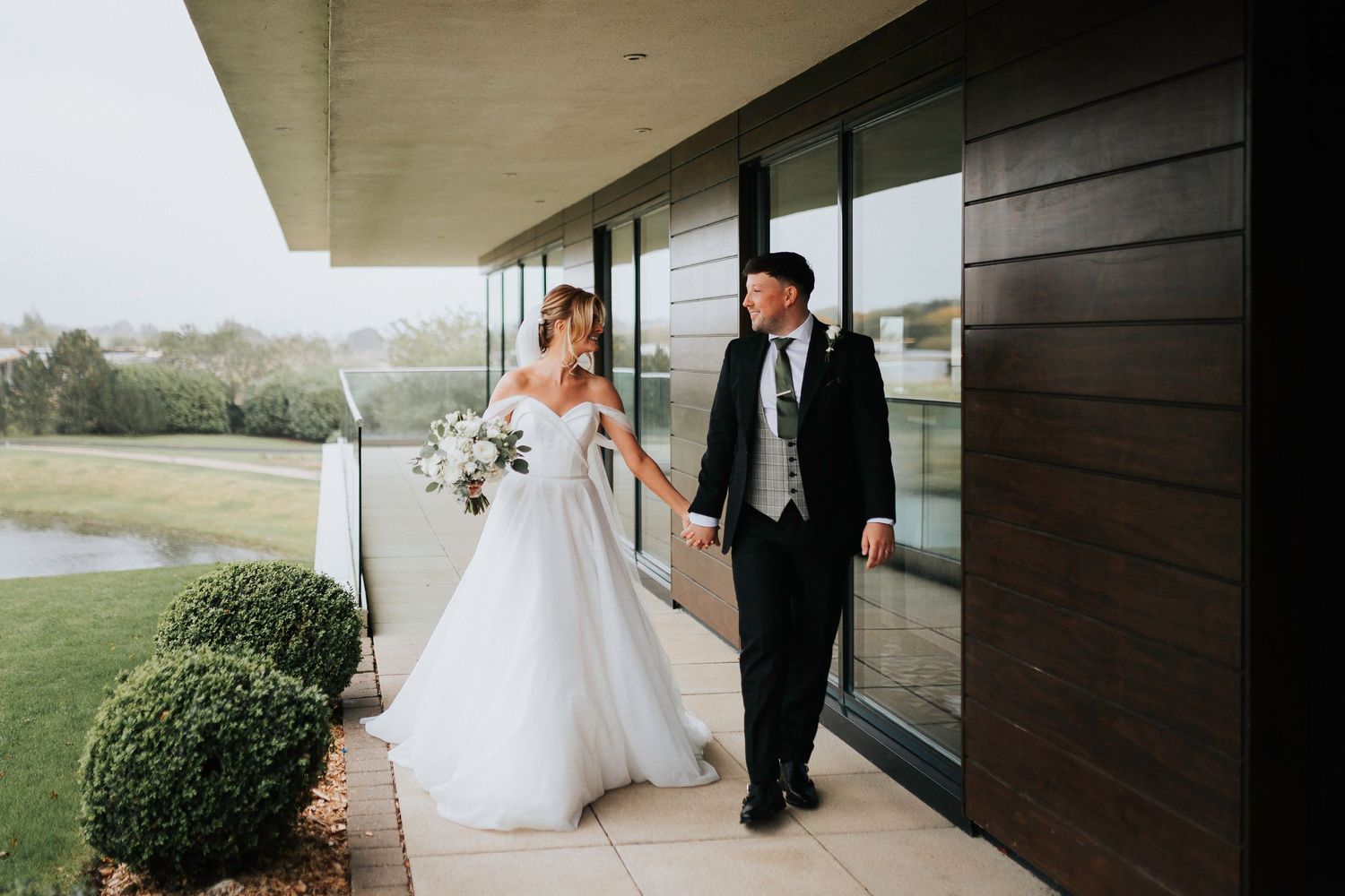 A couple in wedding attire walk together on an outdoor covered porch of a modern building with manicured landscaping.