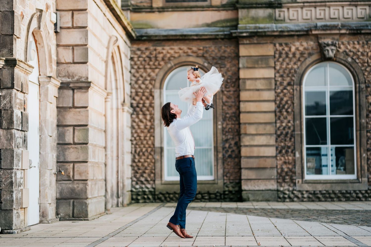 Two people share a loving moment outside an ornate stone building with arched windows.