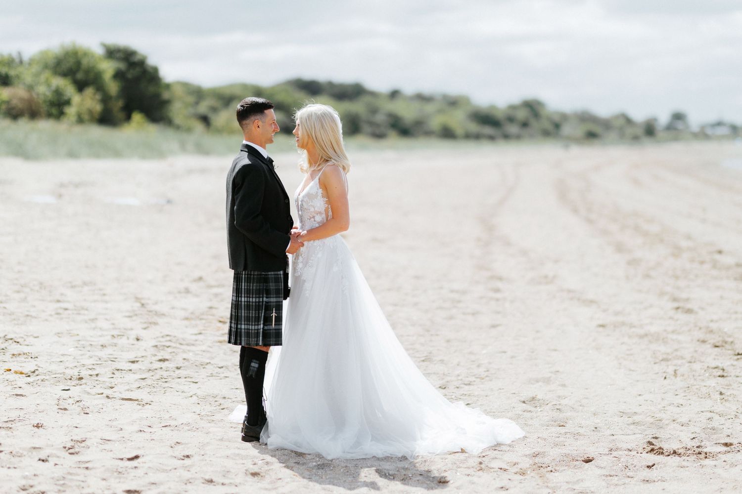 A romantic wedding couple stands together on a sandy beach, with the groom wearing a traditional Scottish kilt.
