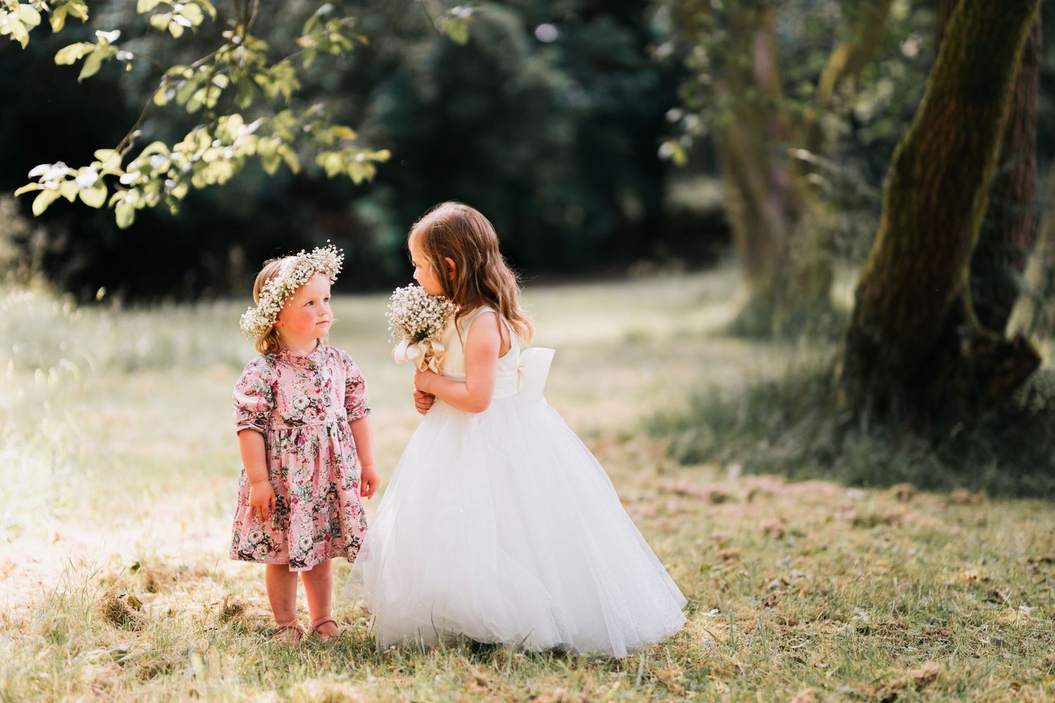 Two young children in dresses stand together in a sunlit garden during a dreamy outdoor photoshoot.