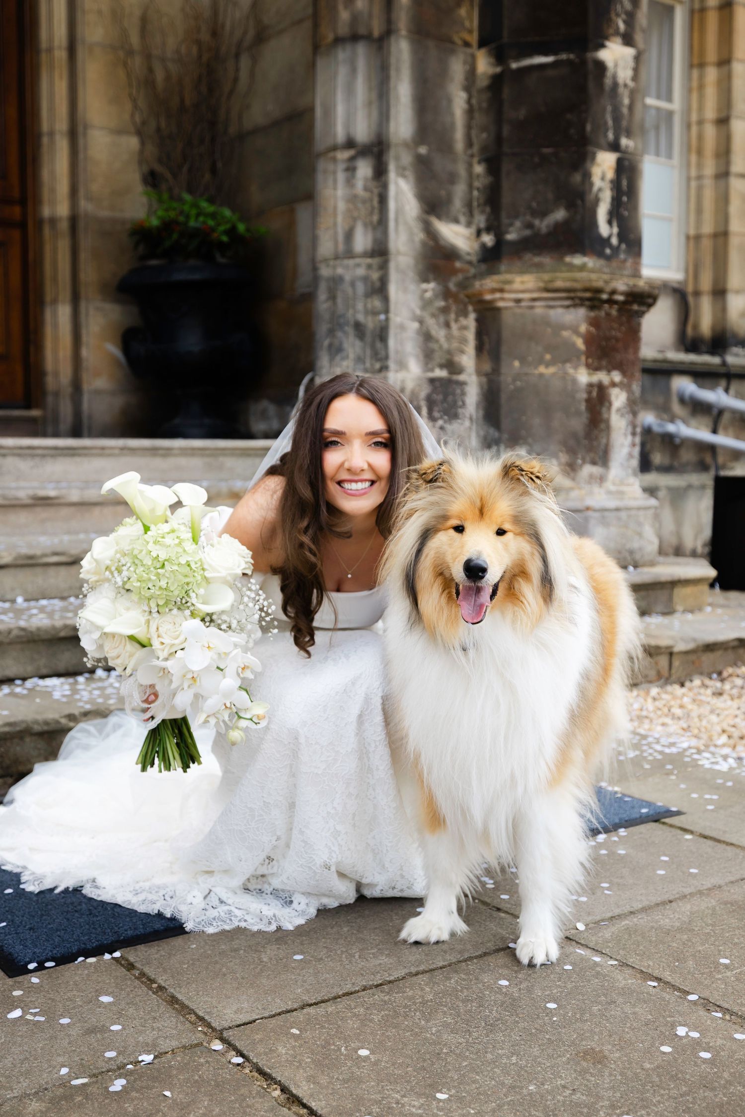 Smiling bride in white gown holding floral bouquet poses with a fluffy Rough Collie dog on stone steps.