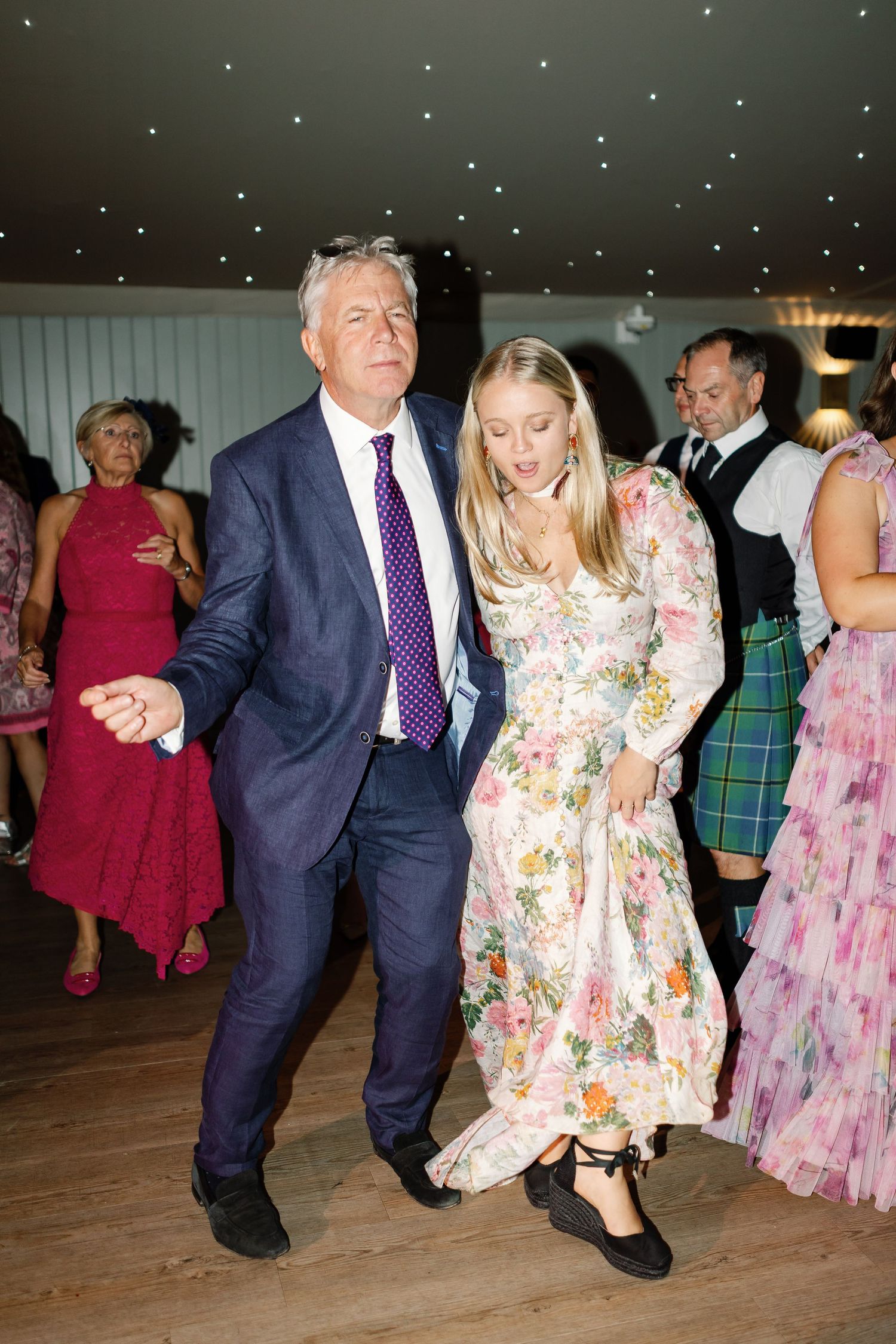 Two people dance together at a formal event, with one wearing a navy suit and purple tie and another in a floral dress.