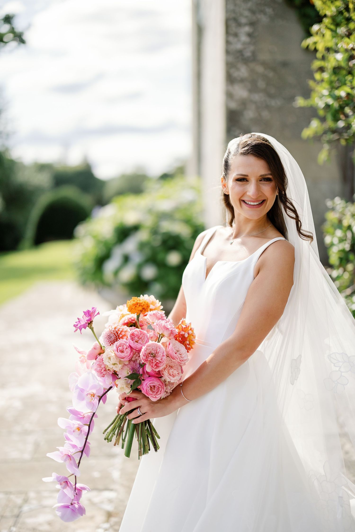 A bride in a classic white wedding gown holds a vibrant pink and orange bouquet in an outdoor garden setting.
