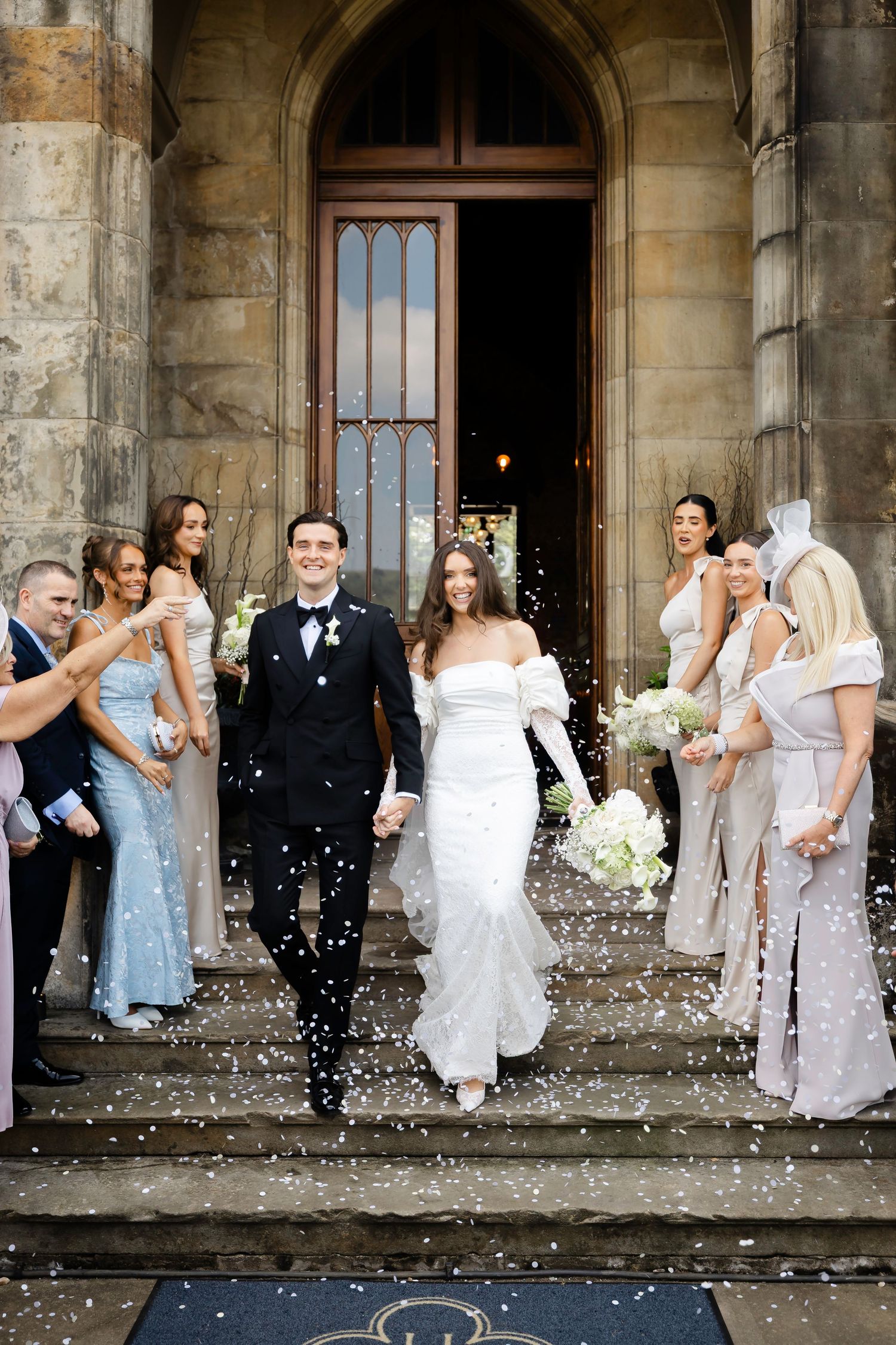 Bride and groom exit stone church amid confetti shower, flanked by bridesmaids in champagne gowns.