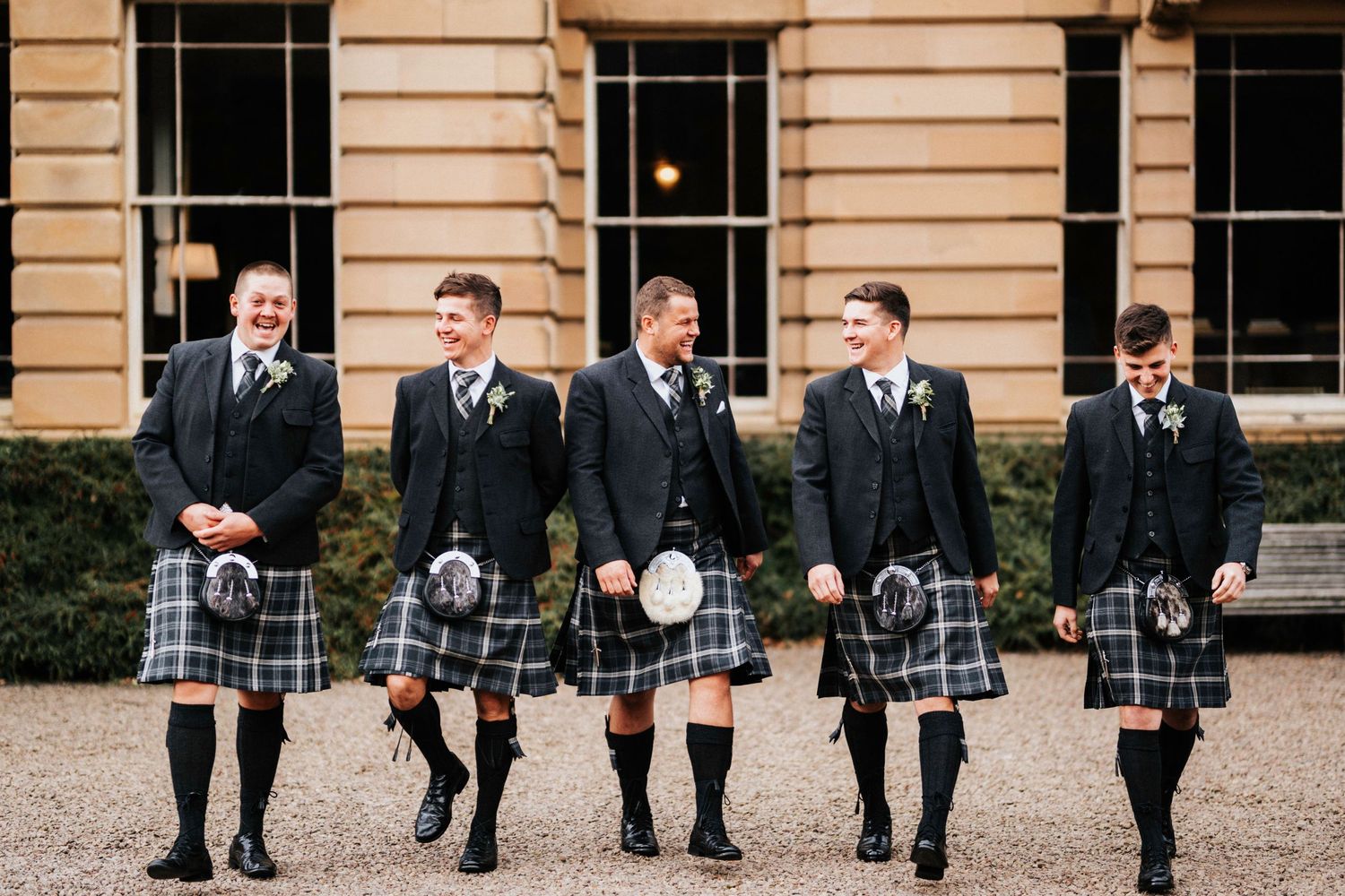 Five men in traditional Scottish kilts and formal jackets stand in a line outside a grand stone building.