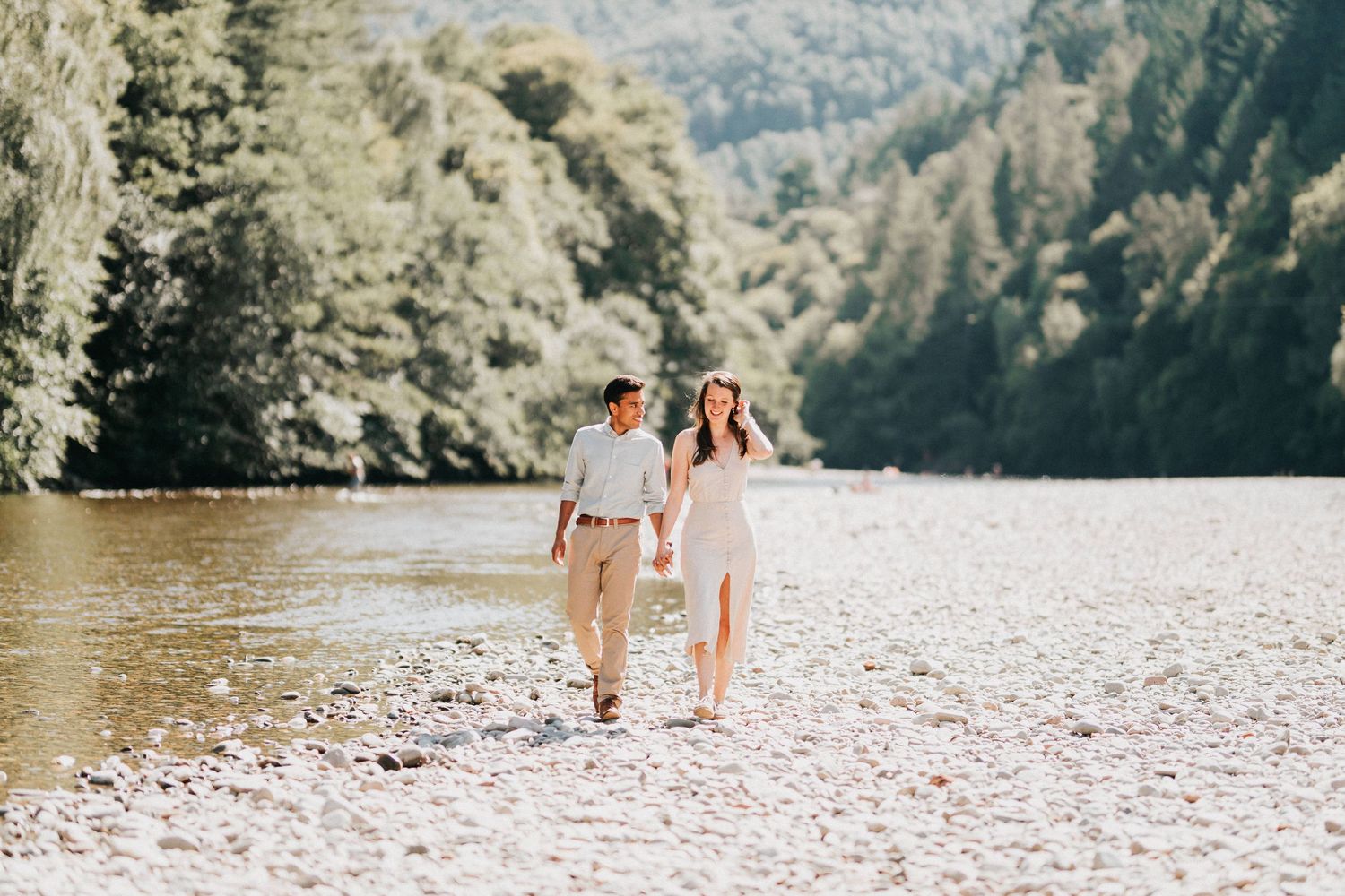A couple walks hand in hand along a scenic rocky riverbank surrounded by lush forest on a sunny day.
