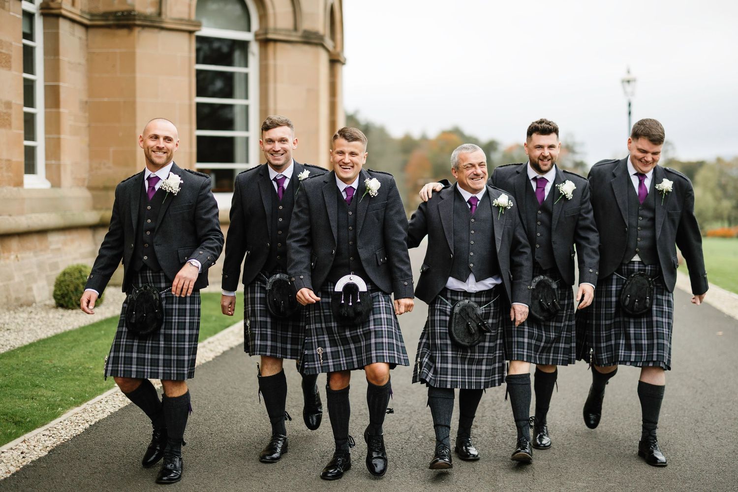 Groomsmen in traditional Scottish kilts and formal attire walk together outside a stone building.
