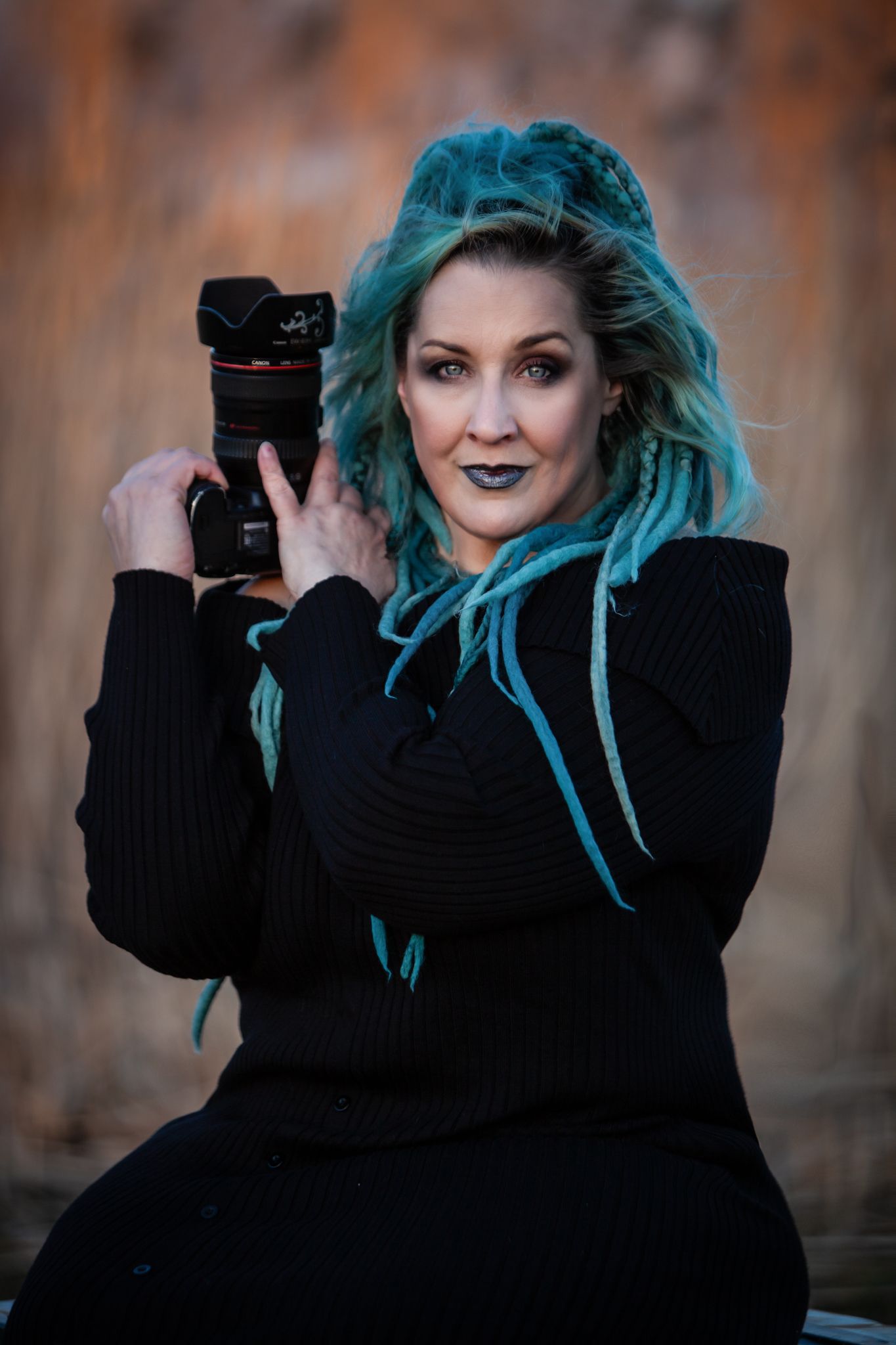 Portrait of someone with vibrant turquoise hair wearing black and holding a camera lens with blurred autumn background.