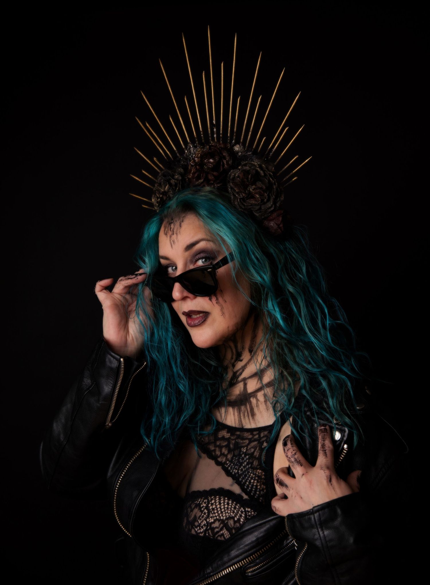 Gothic model in spiked crown and leather jacket with teal hair posing dramatically against black background.