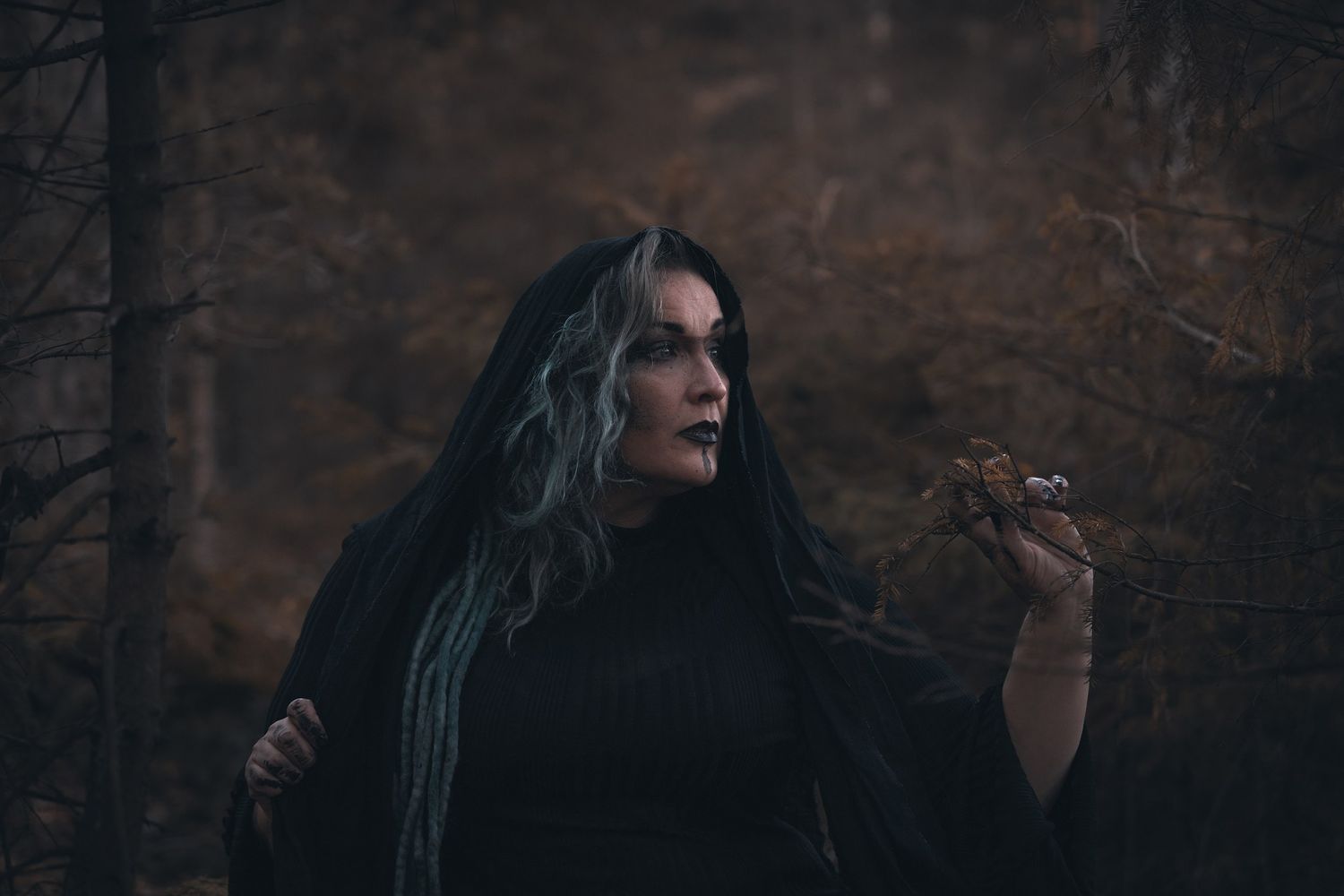 Dark atmospheric portrait outdoors against stone wall showing figure in black with long flowing hair in moody lighting.