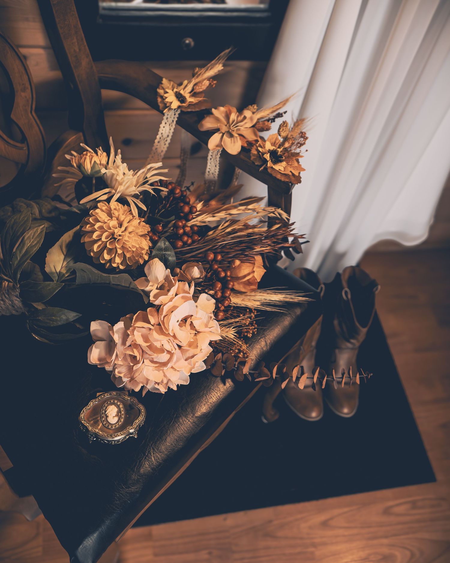 A moody still life arrangement of dried flowers in golden tones displayed on a dark surface near a curtained window.