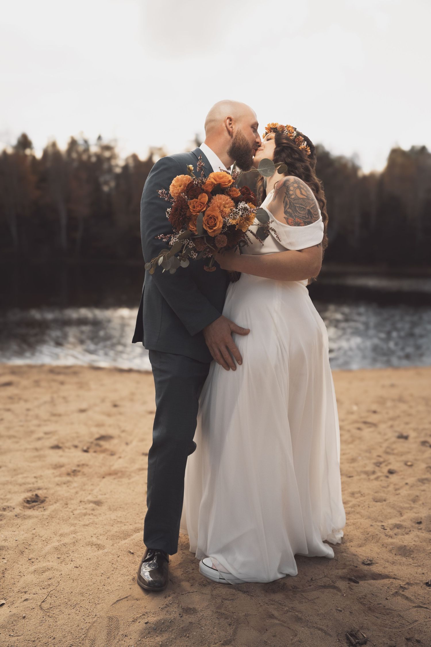 Newlyweds embrace on sandy beach at dusk holding orange floral bouquet with lake and forest in background.