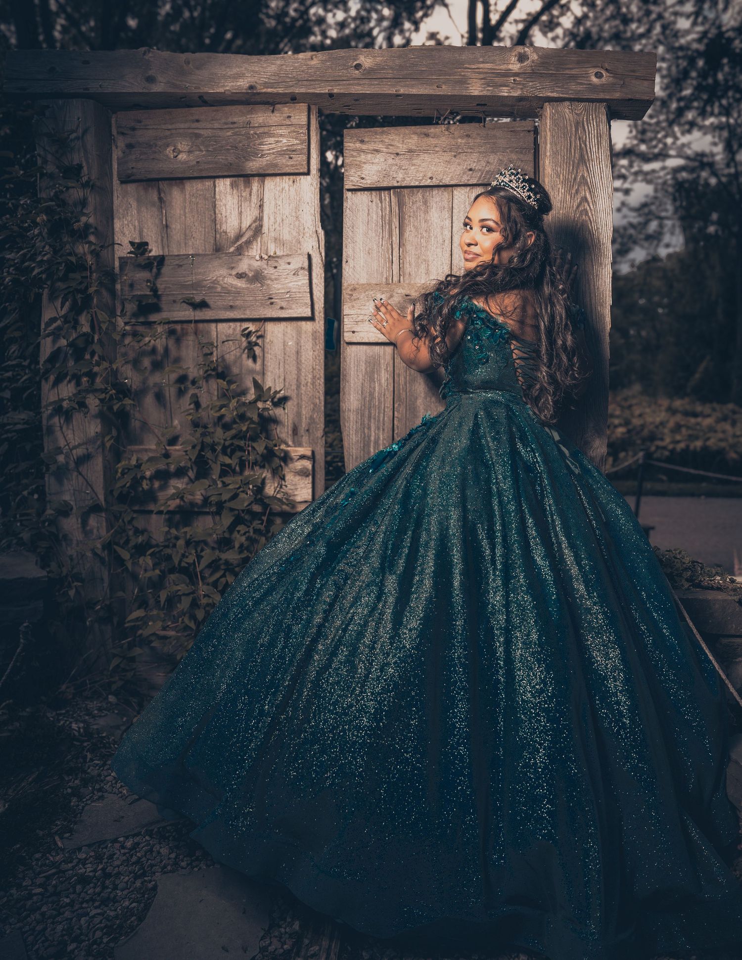 A glamorous sparkly teal ballgown photographed against a rustic wooden door and ivy backdrop in moody lighting.