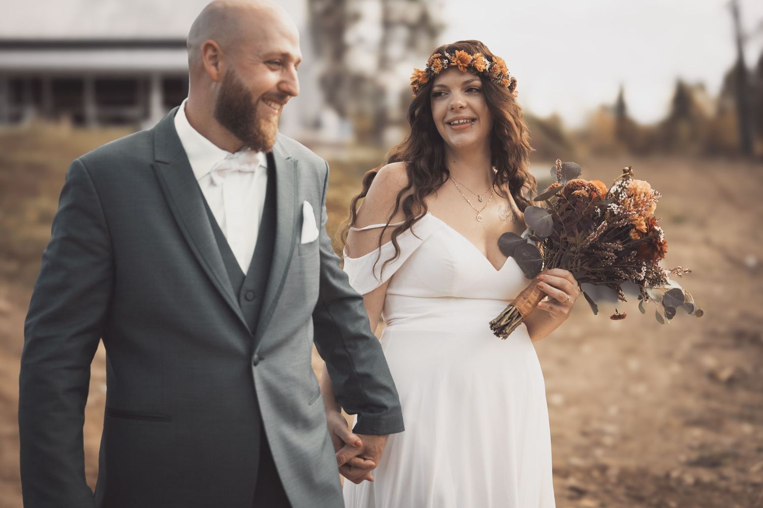 A bride wearing a floral crown and white dress holds a dark bouquet while walking with her groom in a gray suit.